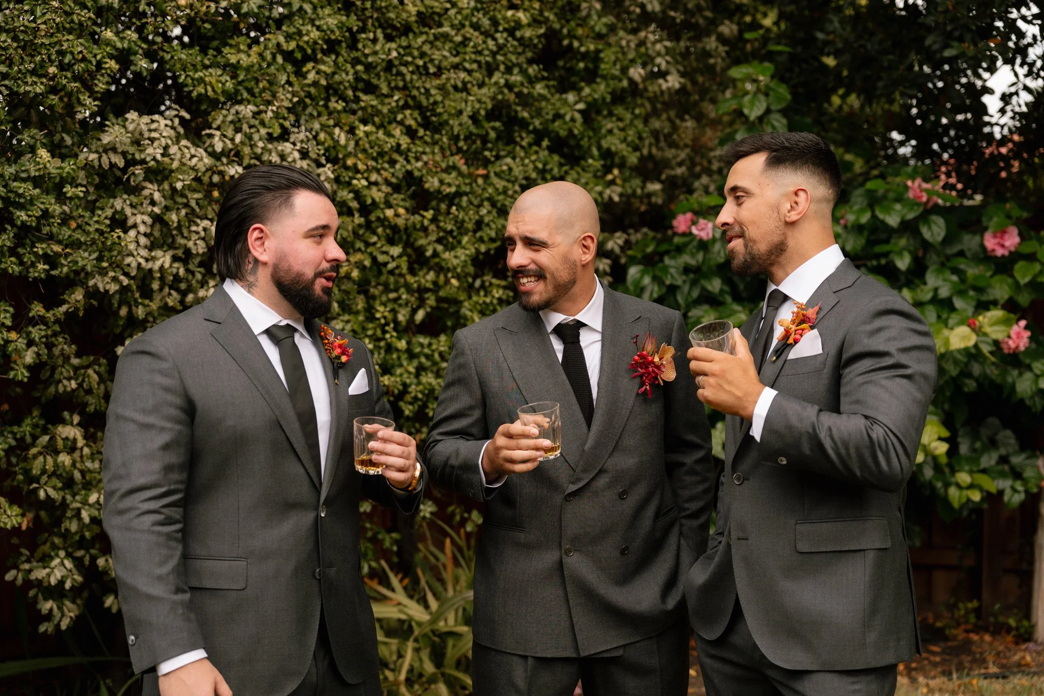 Three men in suits with boutonnieres, holding drinks, talking and smiling outdoors with greenery and flowers in background.