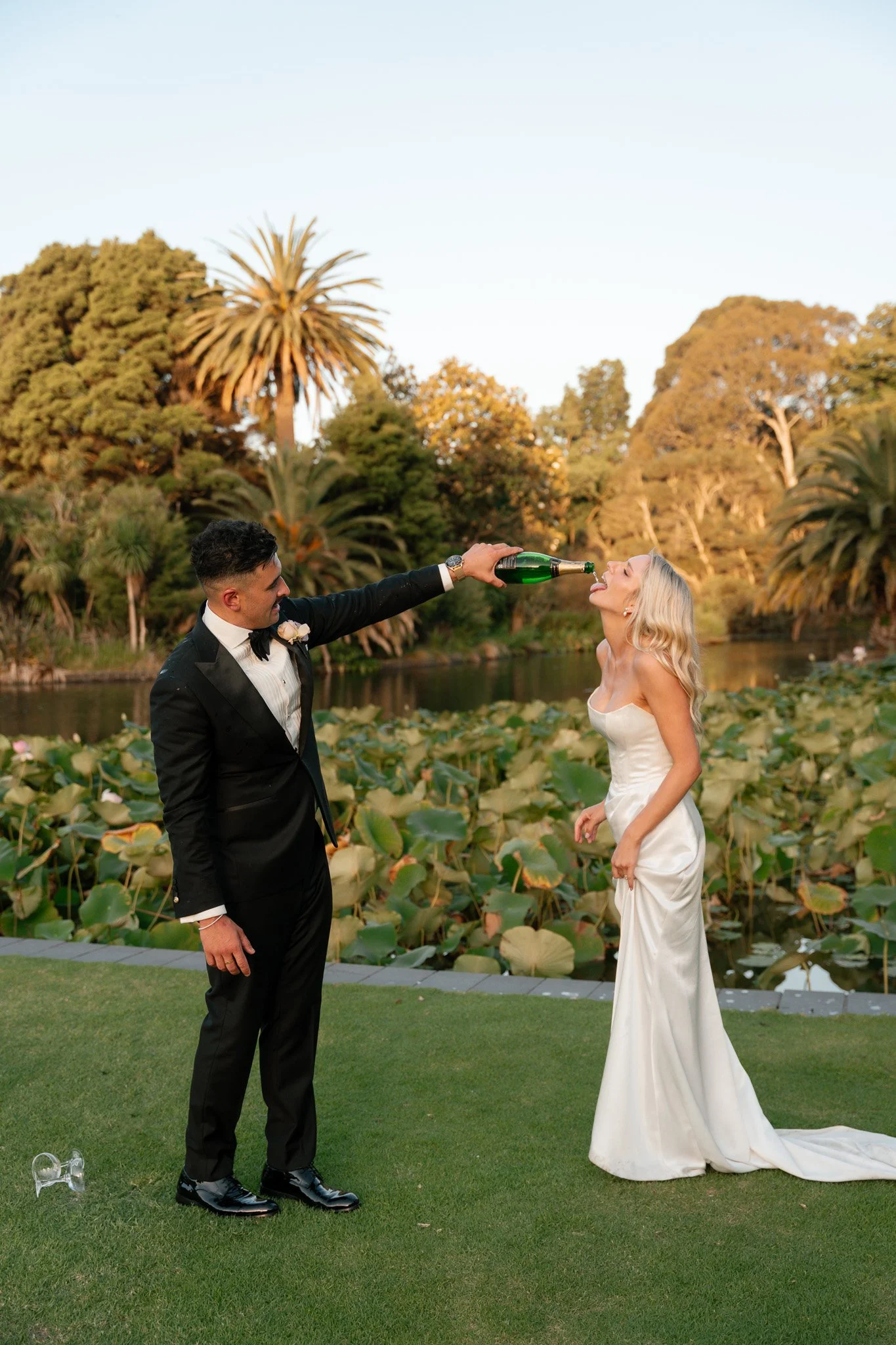 A newlywed couple in wedding attire, the groom in a black tuxedo, pouring champagne into the bride's mouth in a lush outdoor setting with trees, water, and lily pads.