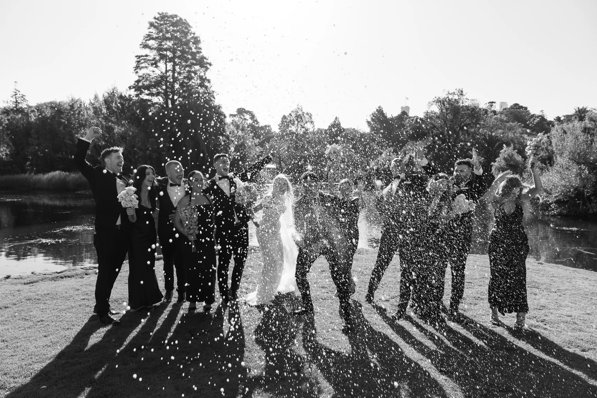 A group of people celebrating, with some dressed in formal attire, standing outdoors near a body of water, with a stream of water splashing around them and sunlight casting shadows.