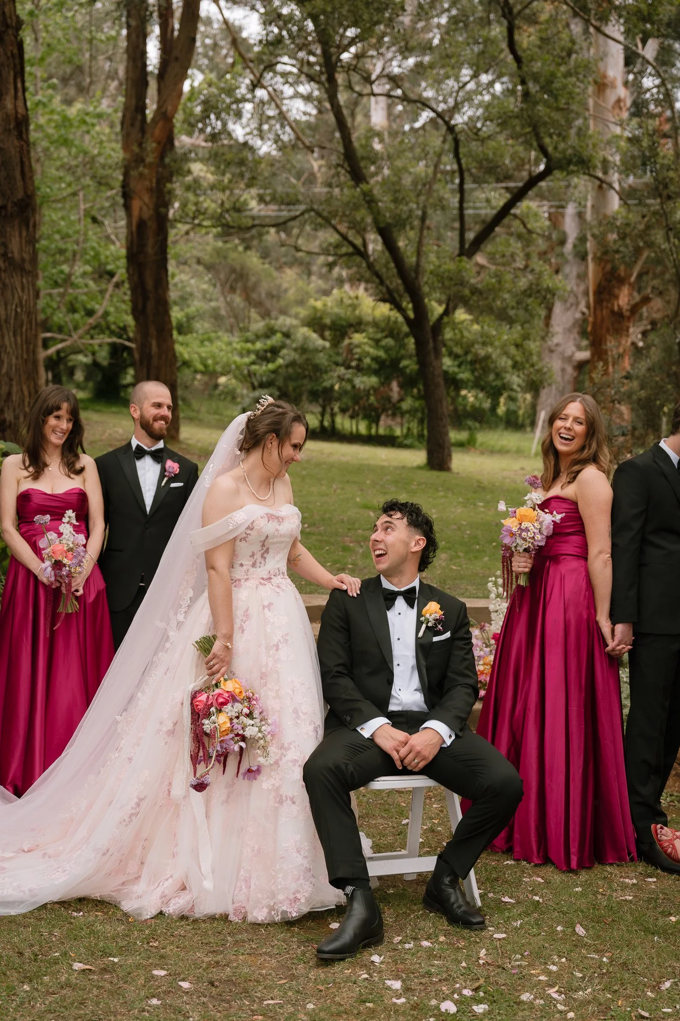 A wedding scene outdoors with the bride and groom surrounded by bridesmaids and groomsmen, all smiling and holding bouquets of flowers.