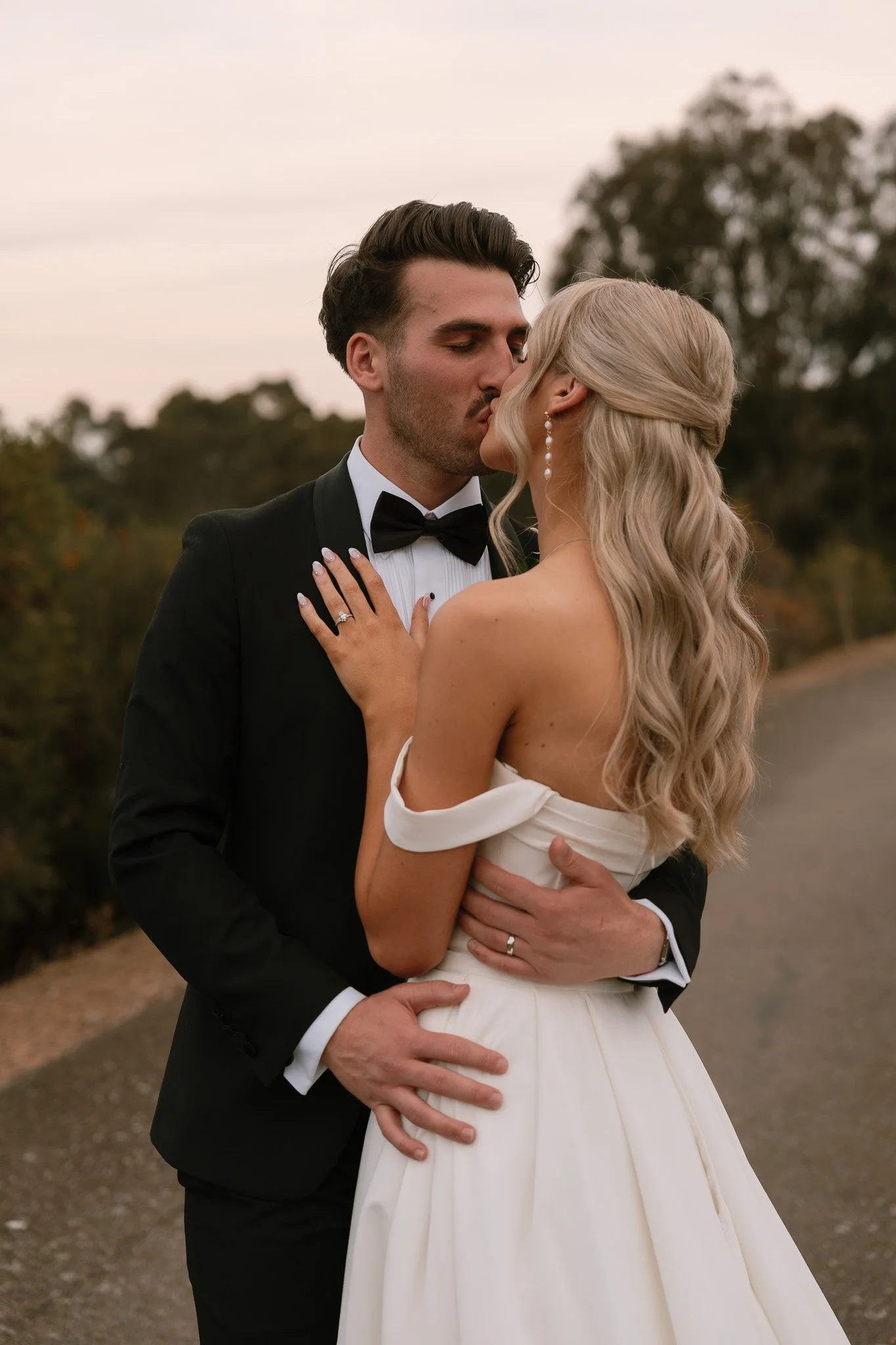 A newlywed couple sharing a kiss outdoors, with the groom in a black tuxedo and the bride in a white off-shoulder wedding gown, during sunset.