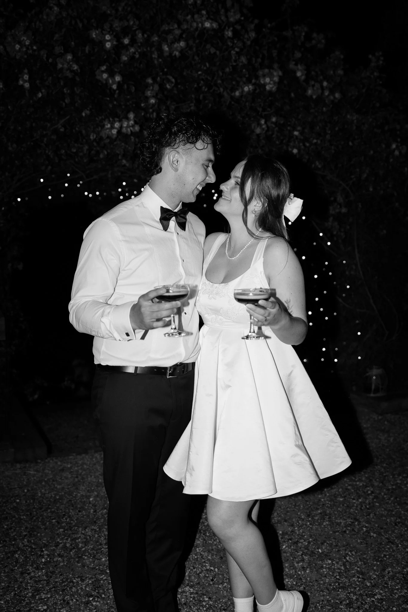 Black and white photo of a couple at a celebration, dressed formally, holding drinks, smiling at each other, with fairy lights in the background.