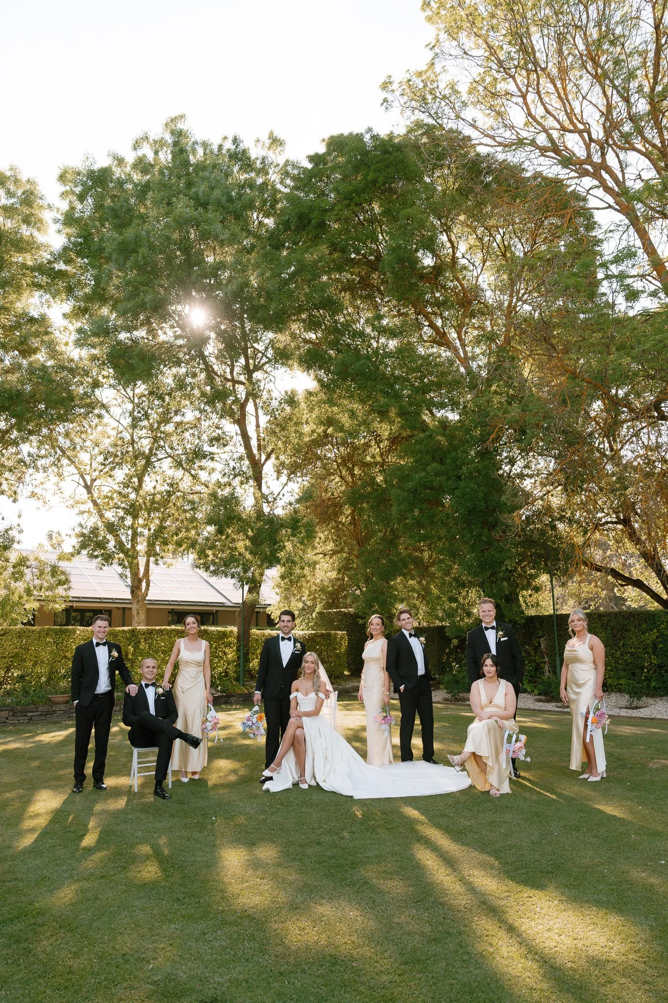 A wedding party standing outdoors on a sunny day, with large trees in the background, including a bride in a white dress, surrounded by bridesmaids in gold dresses and groomsmen in black tuxedos, all smiling and posing.