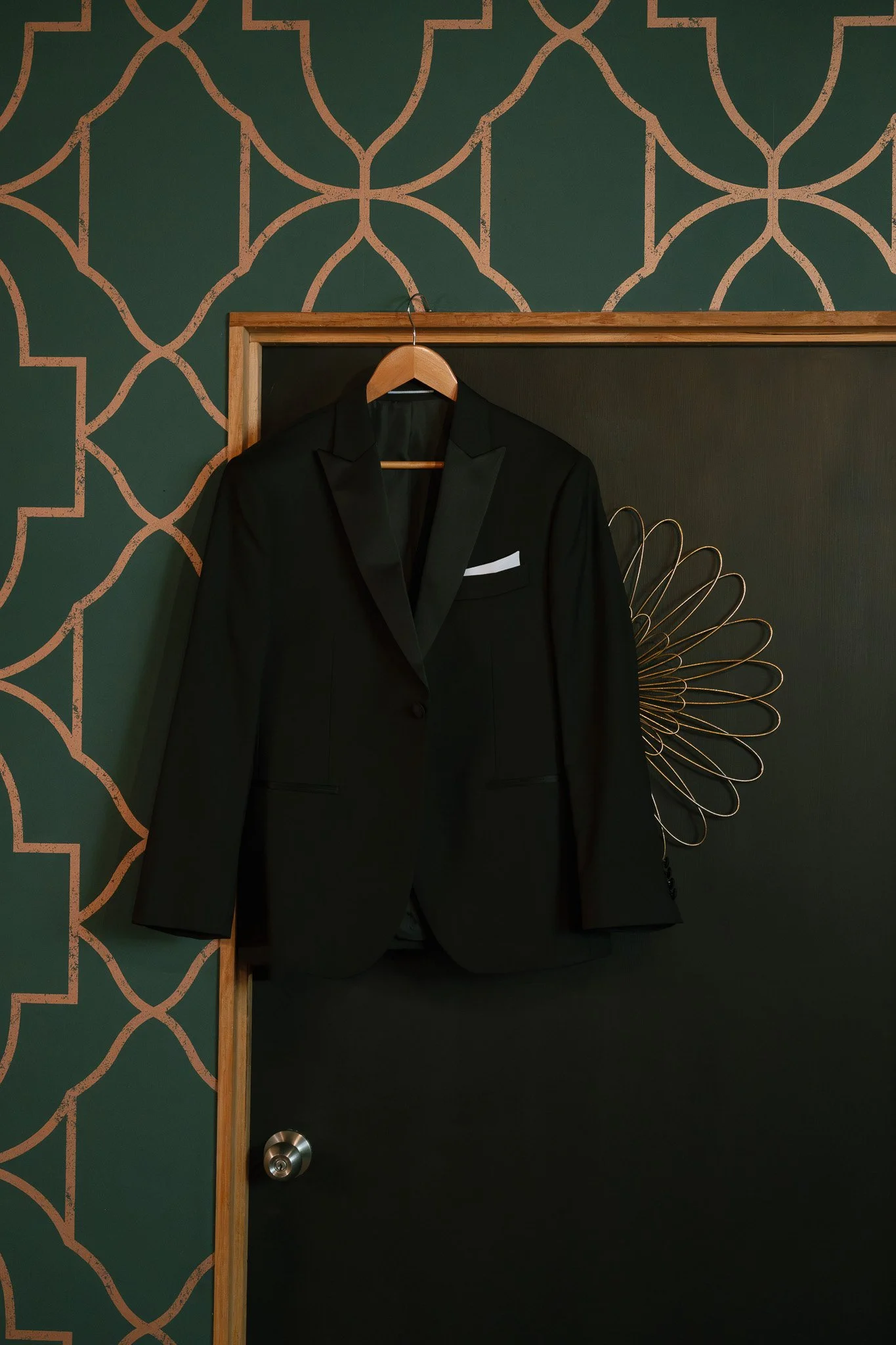 A black tuxedo hanging on a wooden hanger inside a closet, against a dark green and black patterned wall.