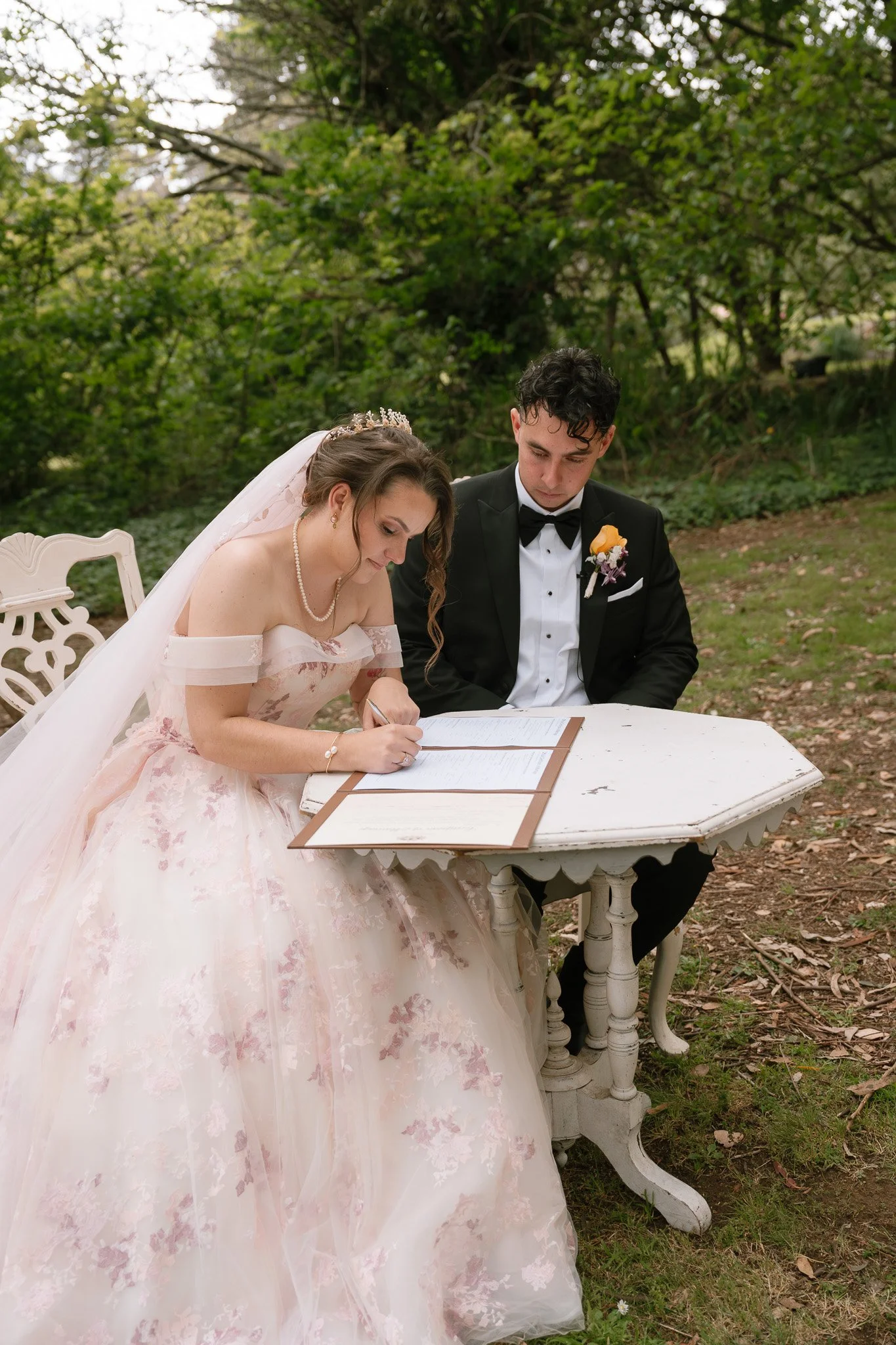 Bride signing a document at outdoor wedding, sitting at a white vintage table with groom looking on, surrounded by trees and greenery.
