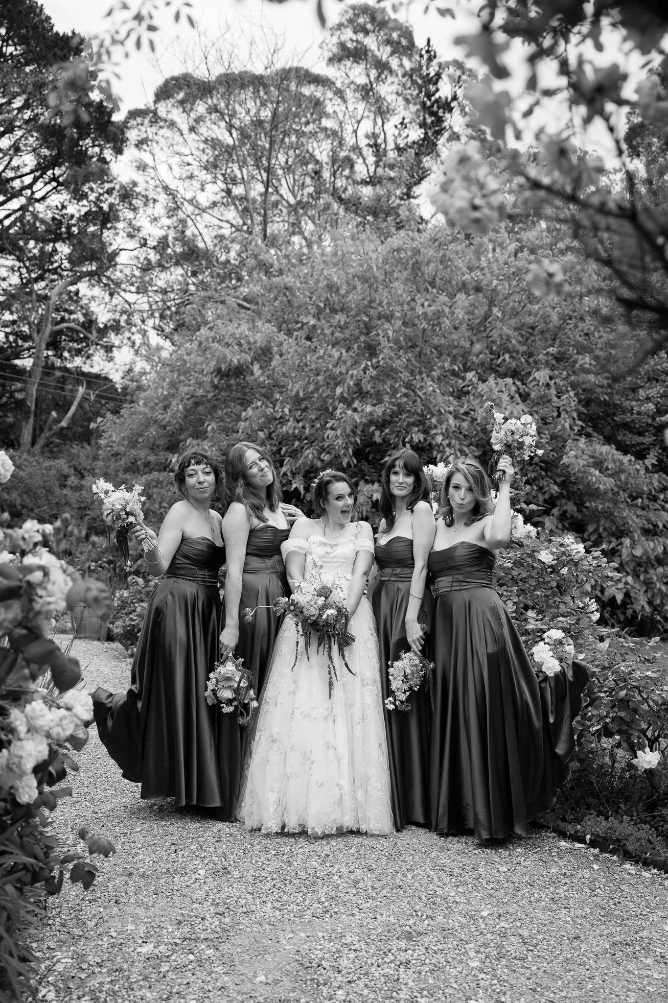 Black and white photo of a bride and four bridesmaids standing outdoors on a gravel path surrounded by trees and bushes, with the bridesmaids wearing matching strapless dresses and holding bouquets, and the bride in a white lace gown holding a bouque