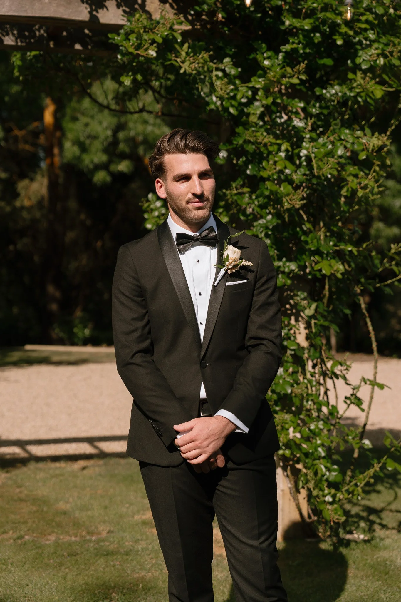 A man in a black tuxedo with a bow tie and boutonniere standing outdoors in front of green foliage.