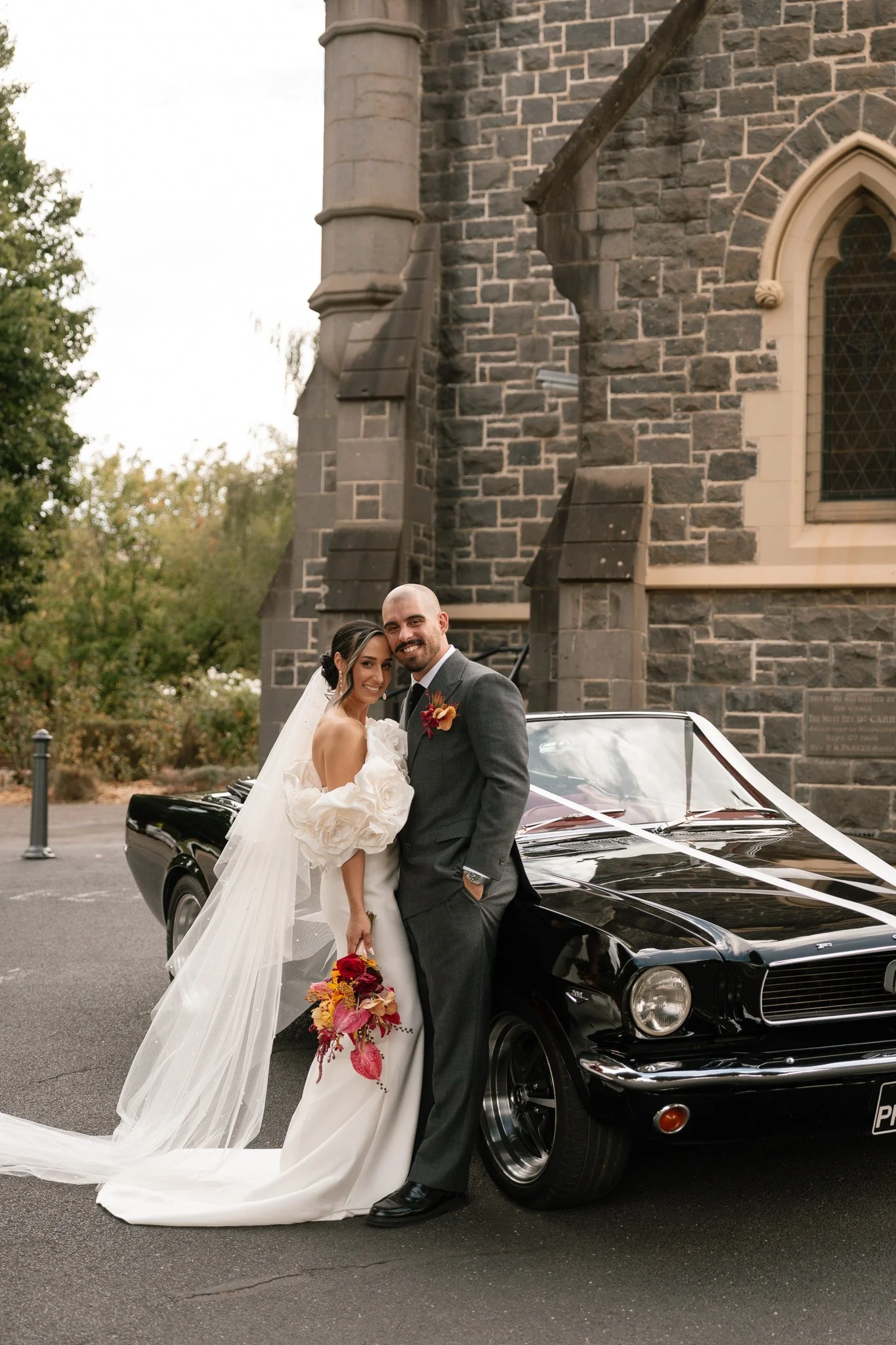 Bride and groom in wedding attire smile and pose beside a black vintage car decorated with white ribbons, in front of a stone church.