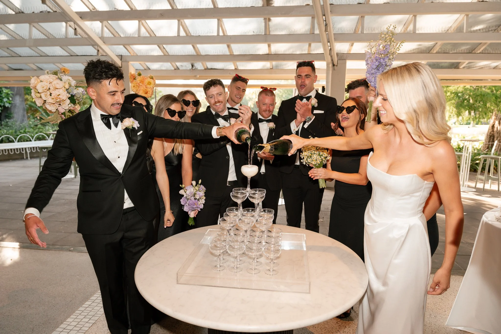 Group of people in formal attire celebrating and pouring champagne into a tower of glasses at a wedding reception.