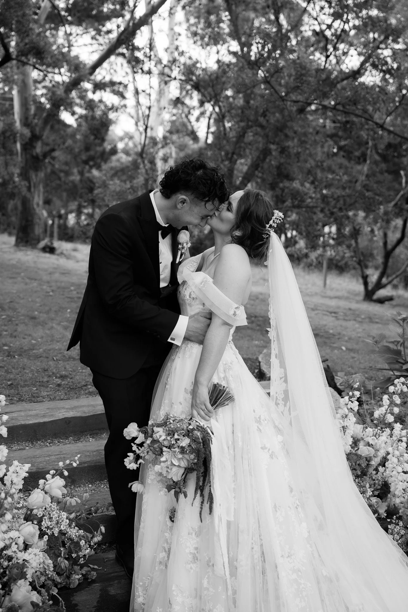 Black and white photo of a bride and groom kissing outside, surrounded by flowers and trees.