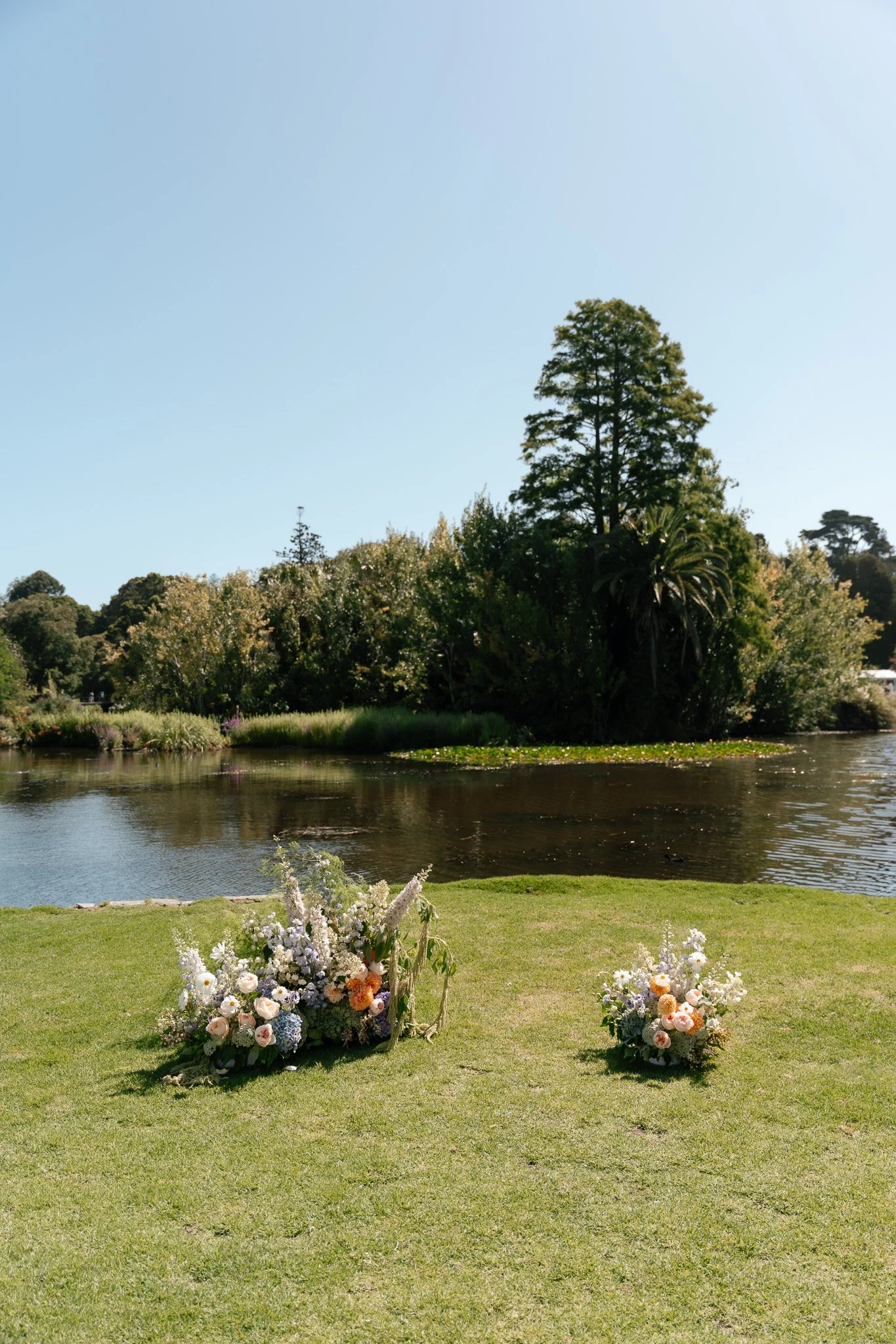 Two flower arrangements on a grassy area near a lake with trees in the background on a sunny day.