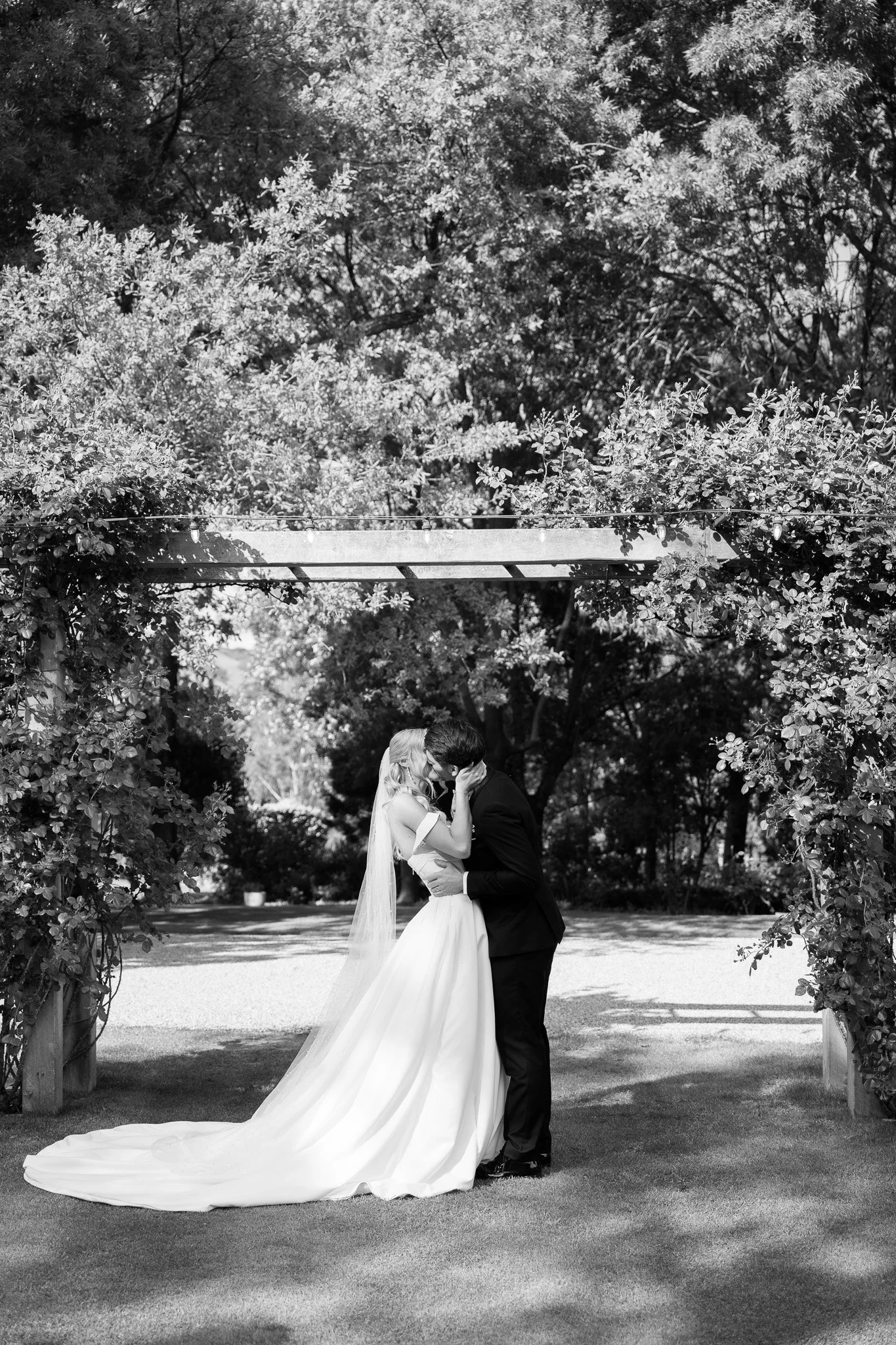 A bride and groom share a kiss under a floral arch outdoors on their wedding day.