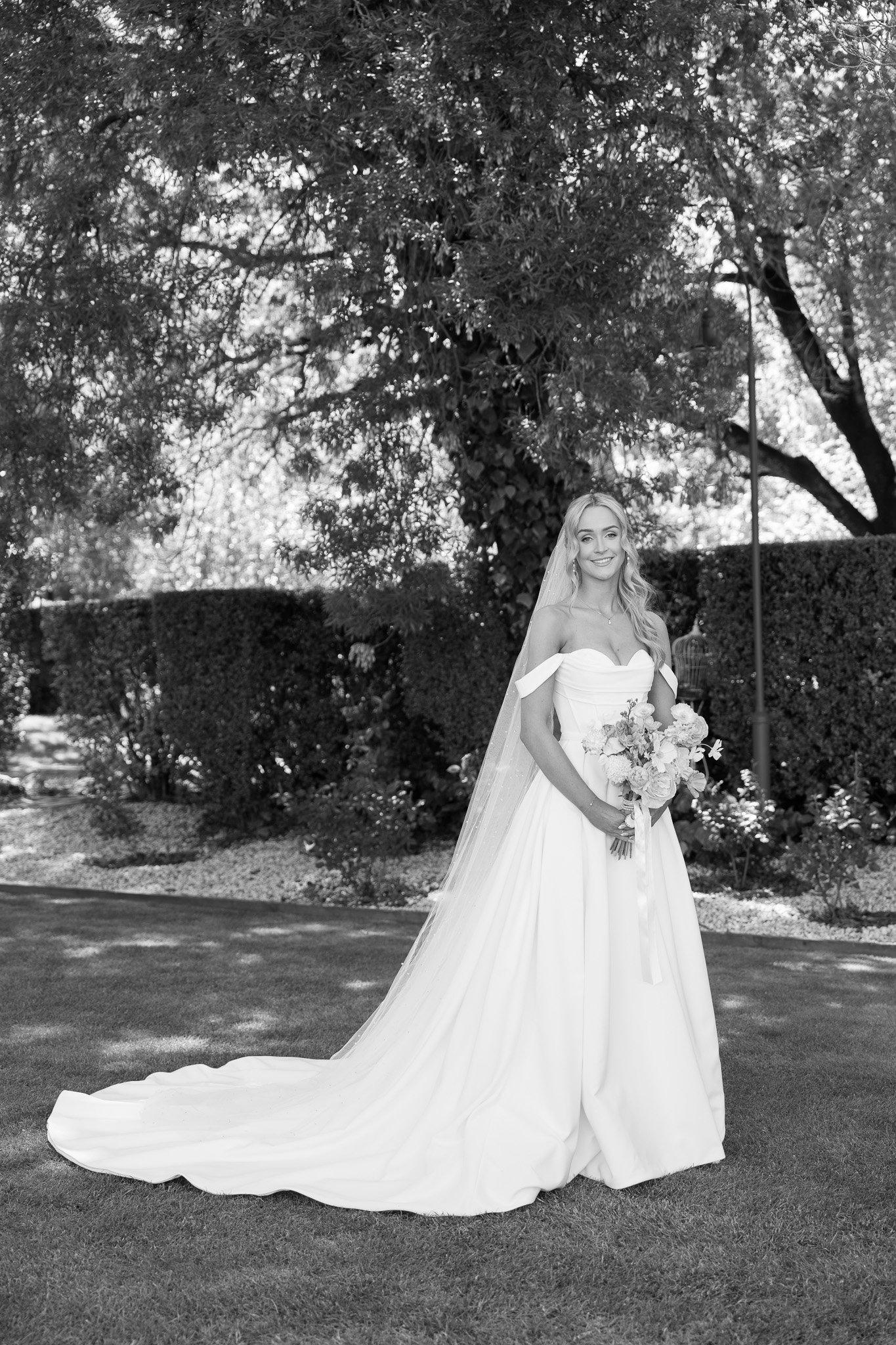 A bride in a white wedding gown holding a bouquet, standing outdoors with trees and bushes in the background.