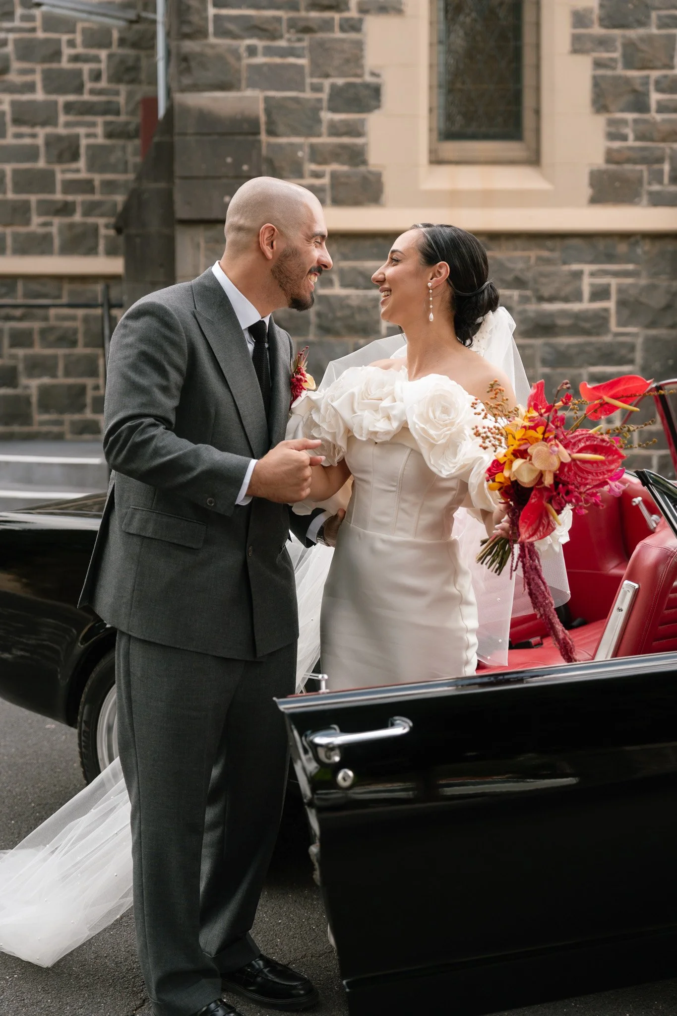 A bride and groom smiling and holding hands beside a black vintage car with a red interior, set against a stone building backdrop.