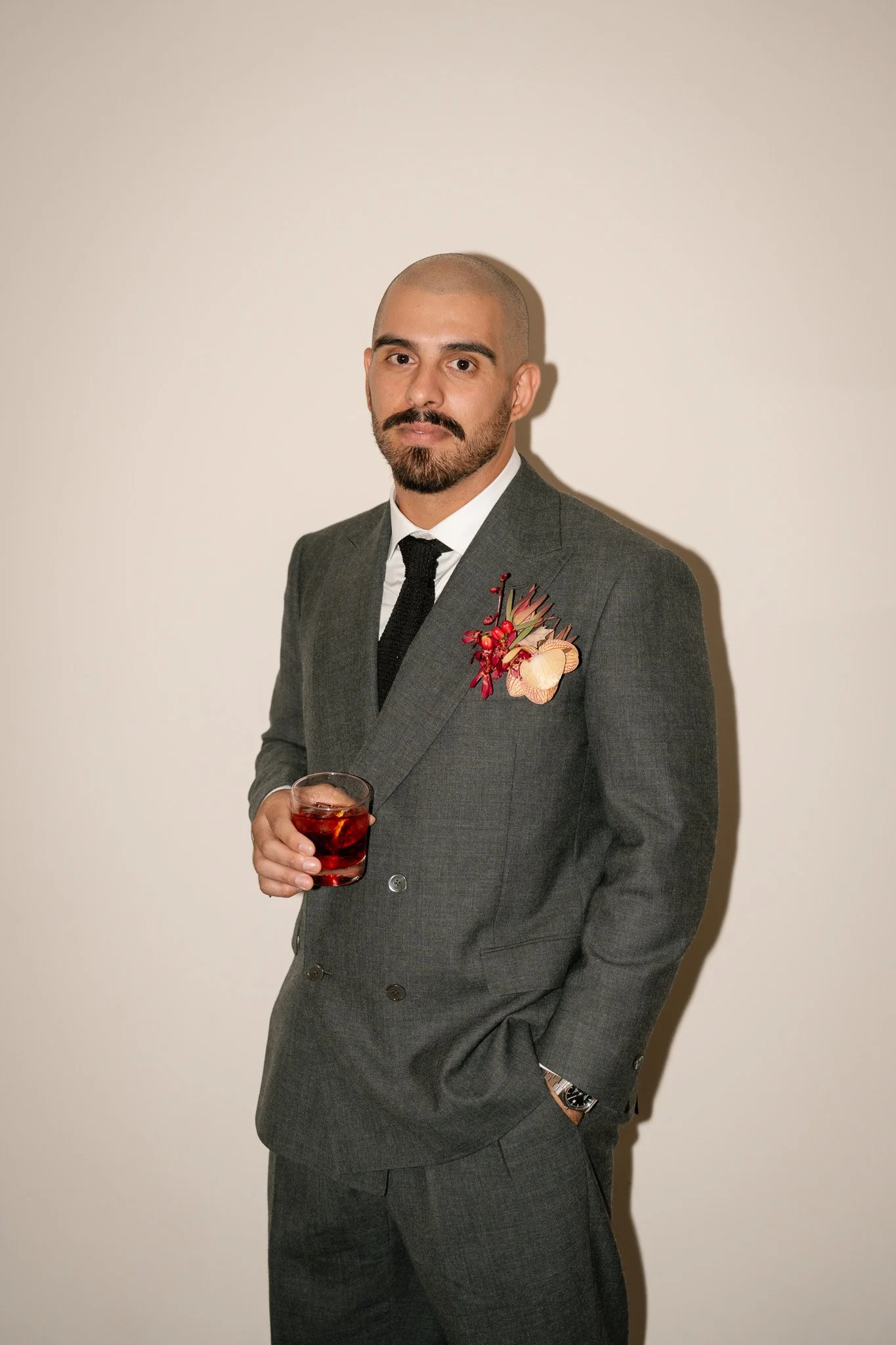 A man in a gray suit holding a glass of red drink, standing against a plain white wall.