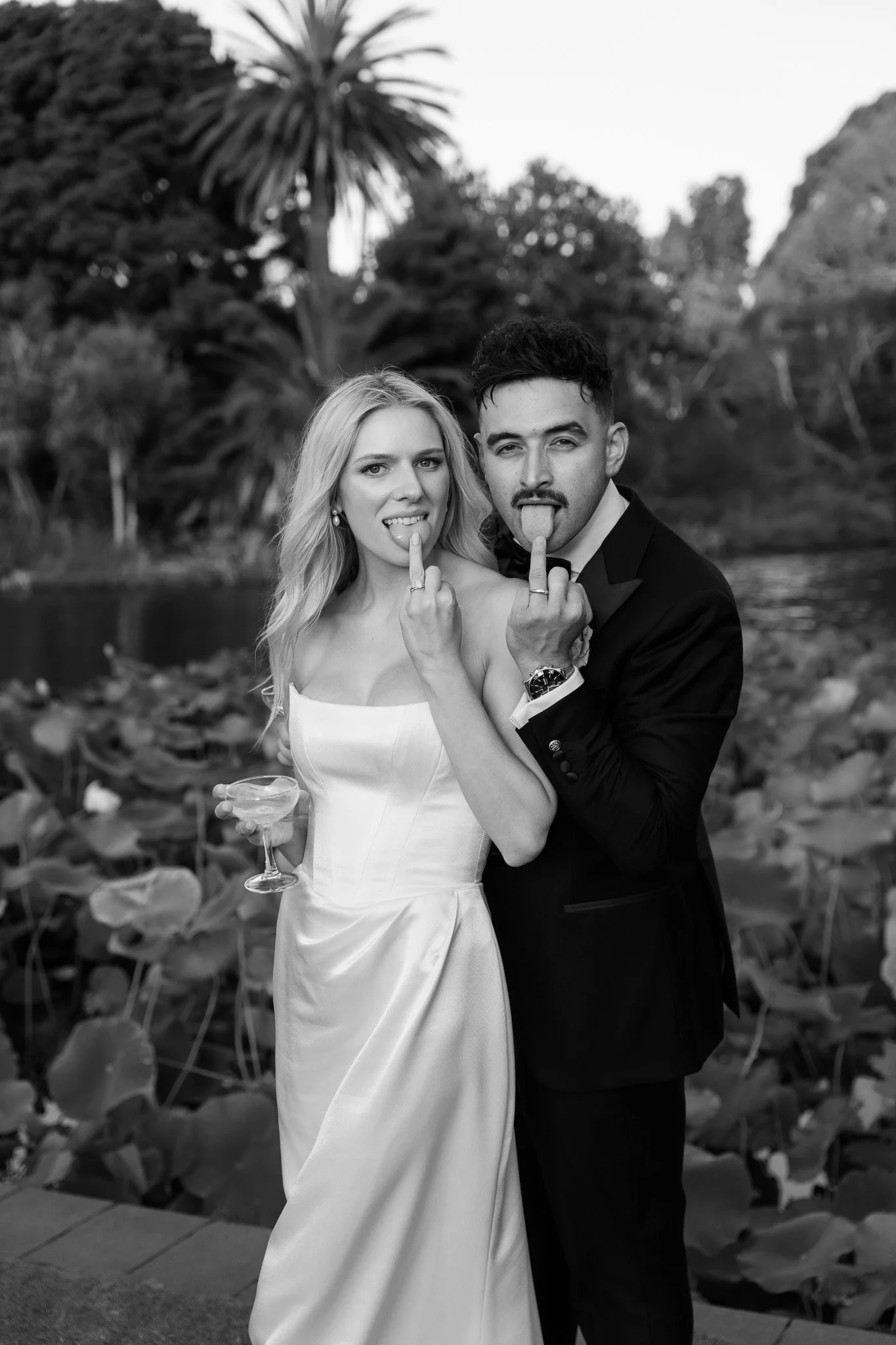 A black and white photo of a woman in a wedding dress and a man in a tuxedo making silly faces, with their tongues out and middle fingers raised. They are standing outdoors near a body of water with lush trees and plants in the background.