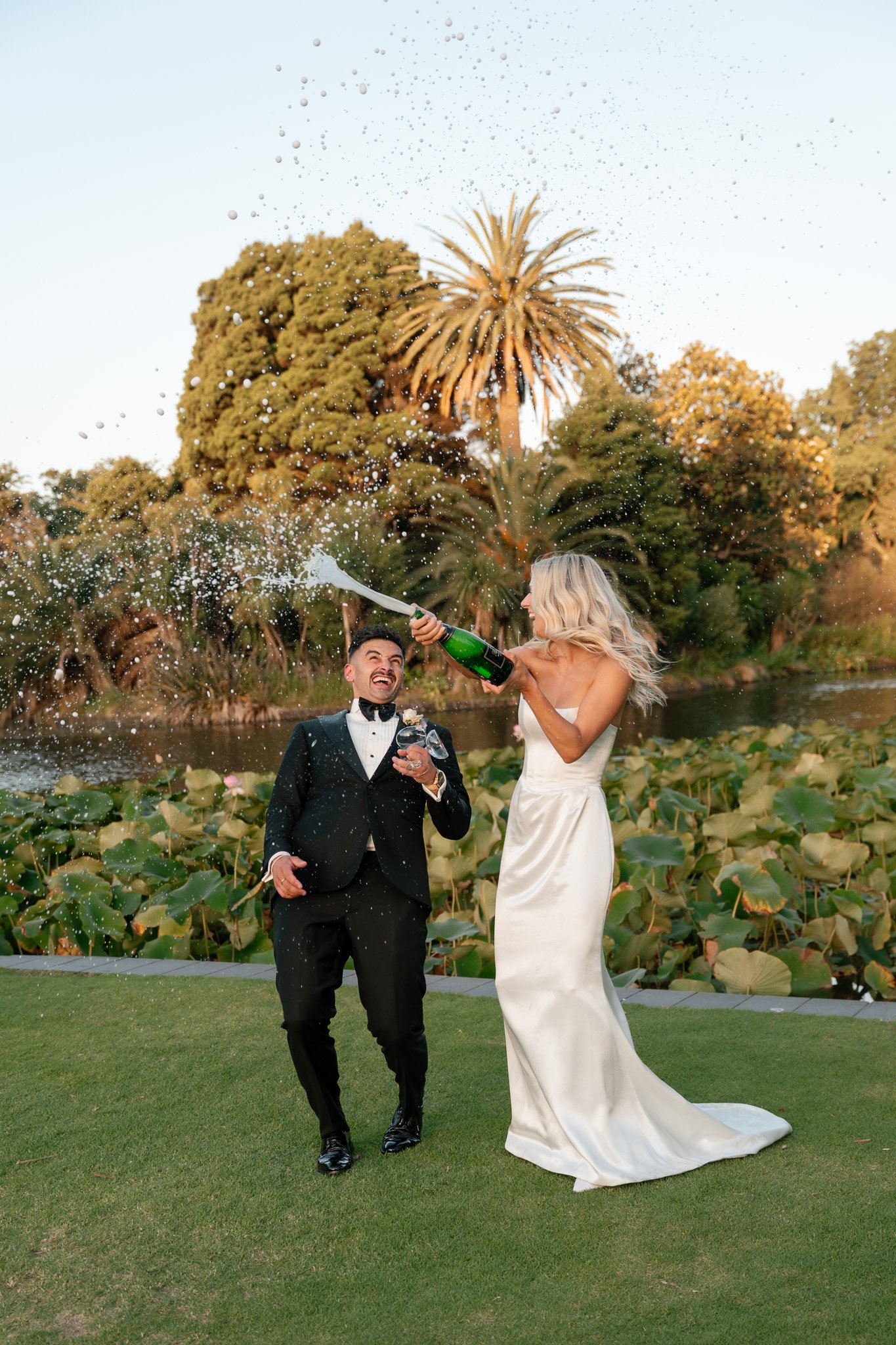 A bride and groom celebrating outdoors by a pond, with the bride spraying champagne on the groom's face.