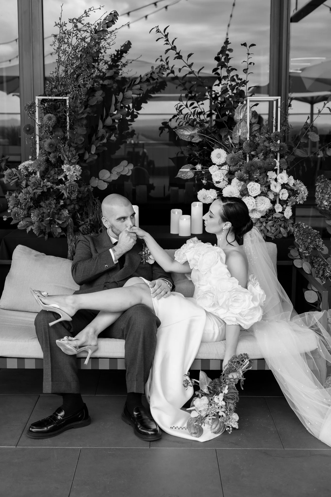 A wedding scene with a bride and groom sitting on a couch, the groom kissing the bride's hand. The bride is dressed in a wedding gown with a veil and bouquet, and the groom is wearing a suit. There are candles and floral arrangements behind them.