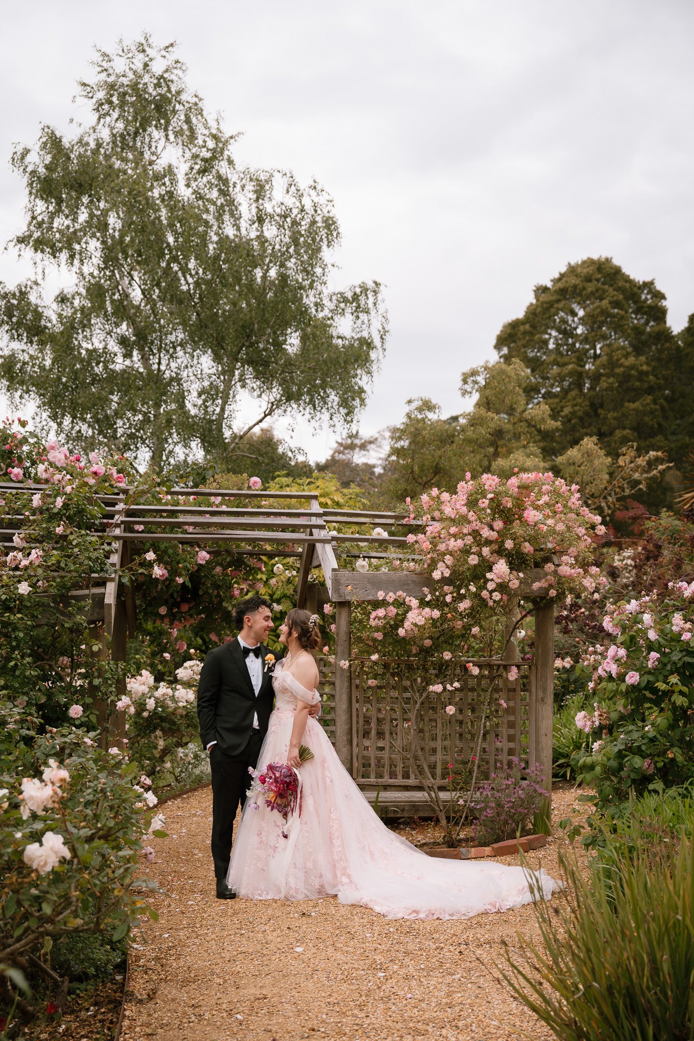 A bride and groom standing together in a garden with pink flowers and trees, sharing a moment on their wedding day.