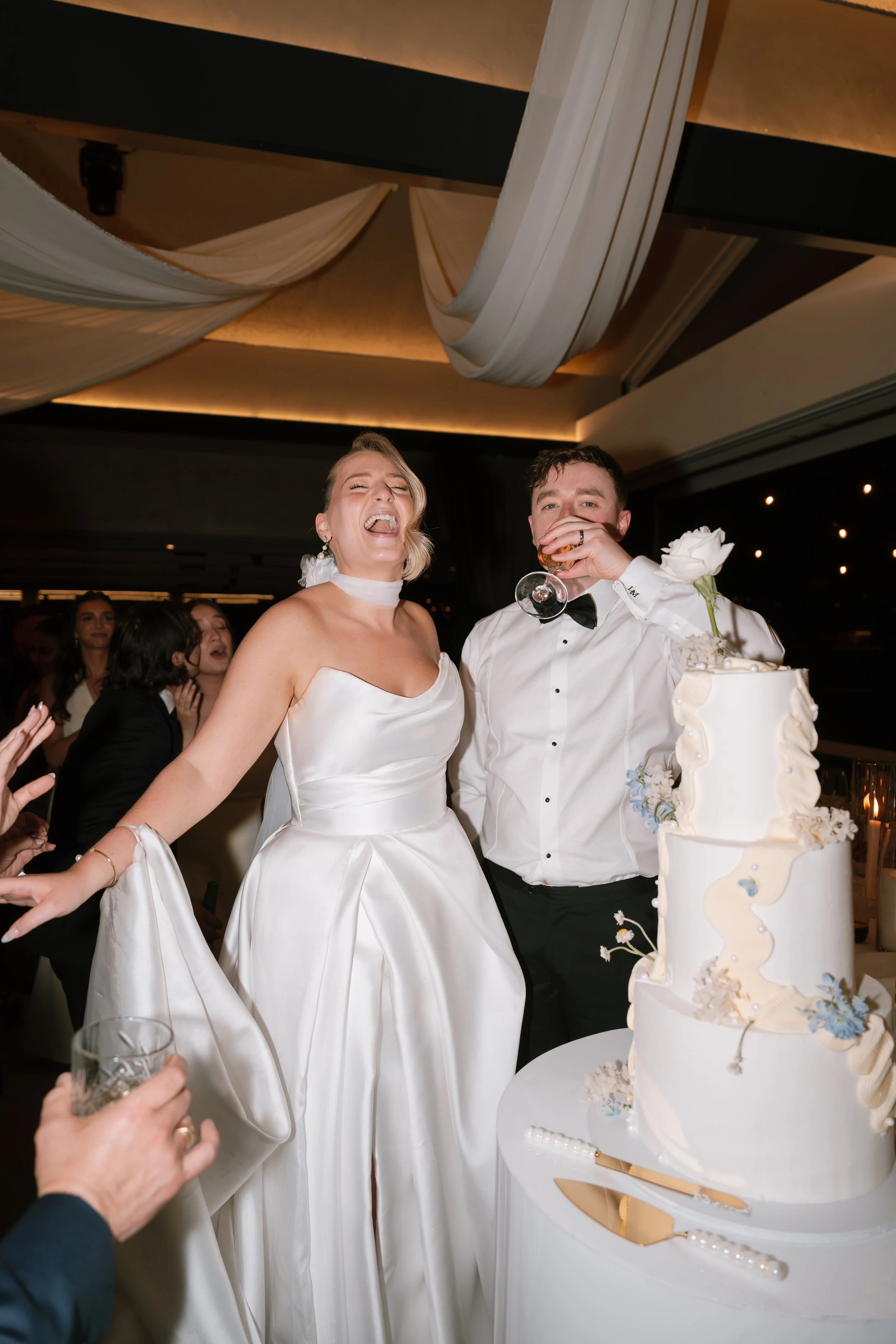 A bride and groom smiling and celebrating at their wedding reception, with a wedding cake in front of them and guests around them.