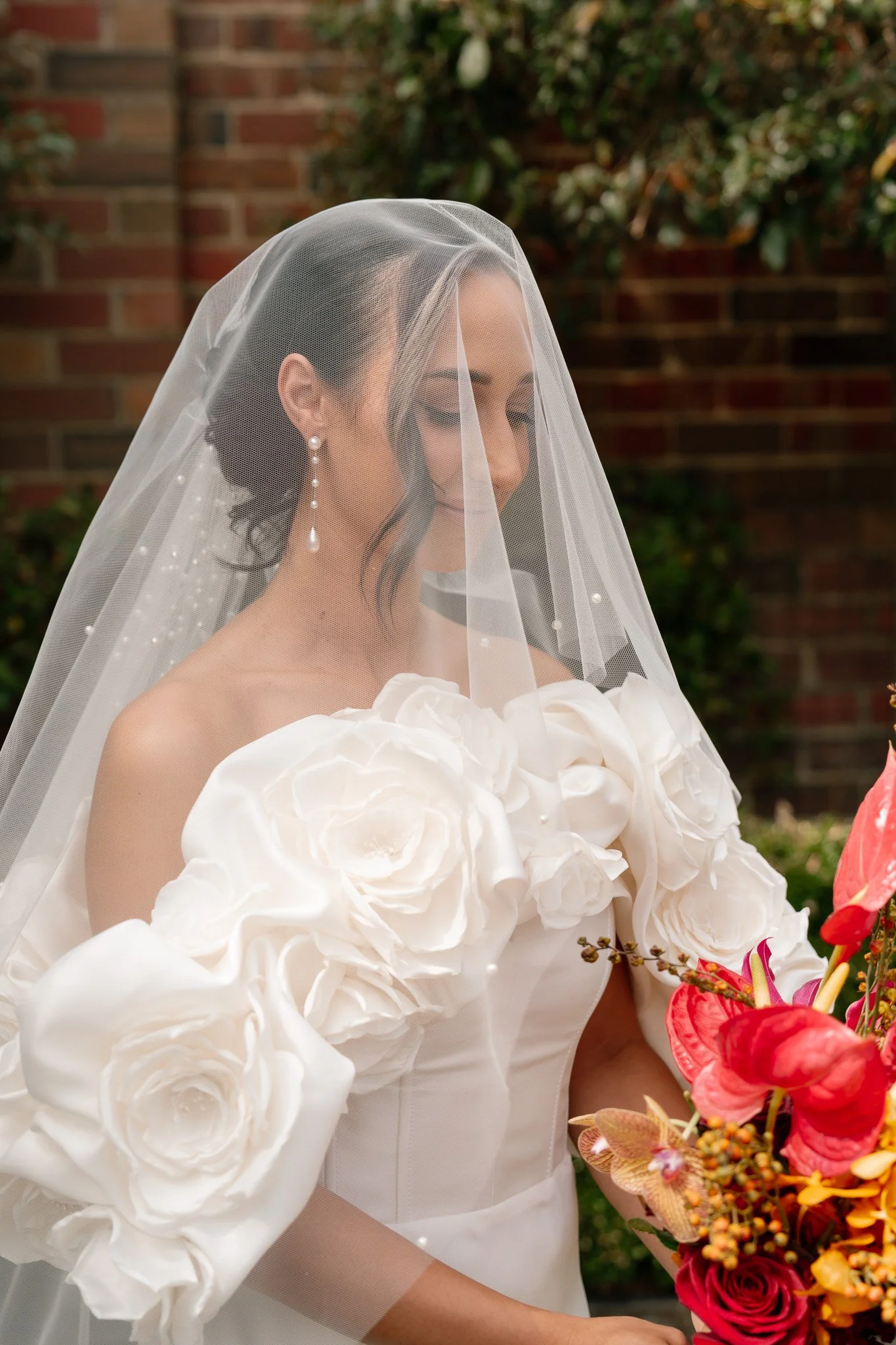 Brunette woman in a wedding dress with large white floral accents, wearing a sheer veil and pearl earrings, holding a bouquet of colorful flowers, standing outdoors in front of a brick wall and greenery.