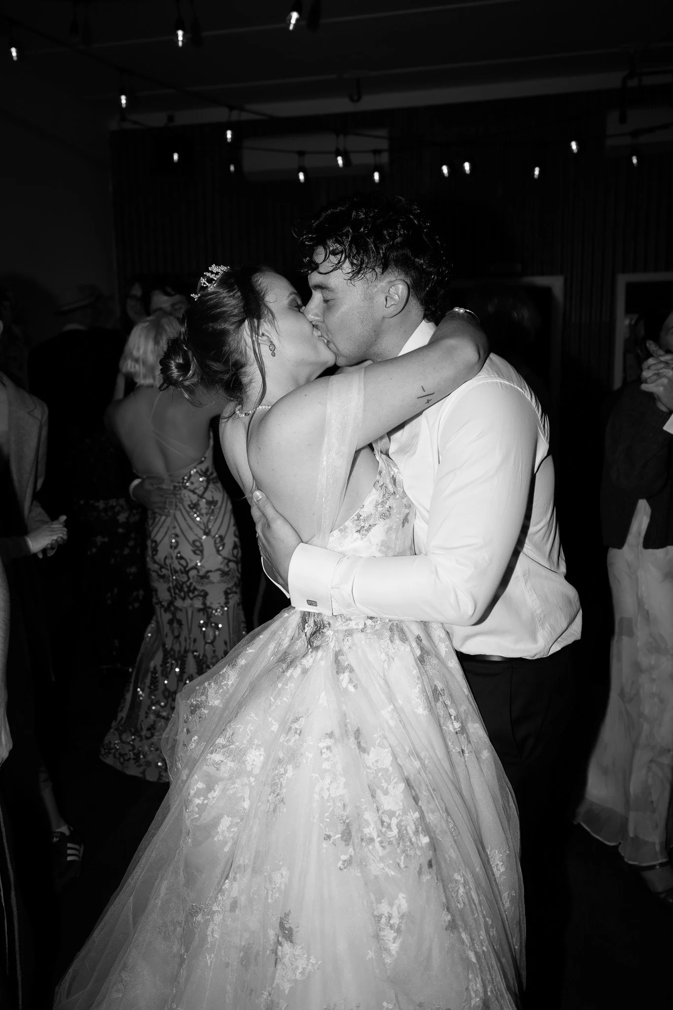 Black and white photo of a couple dancing and kissing at a wedding reception, with guests in the background.