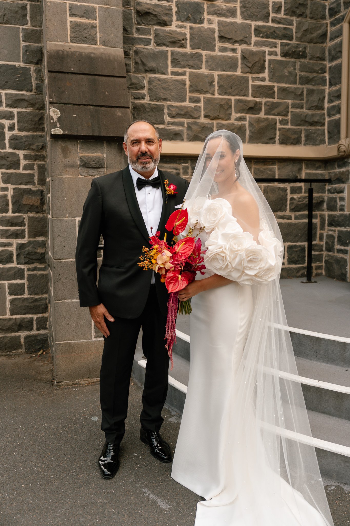A bride in a white wedding gown holding a colorful bouquet, standing next to a man in a black tuxedo with a bow tie, in front of a stone wall.