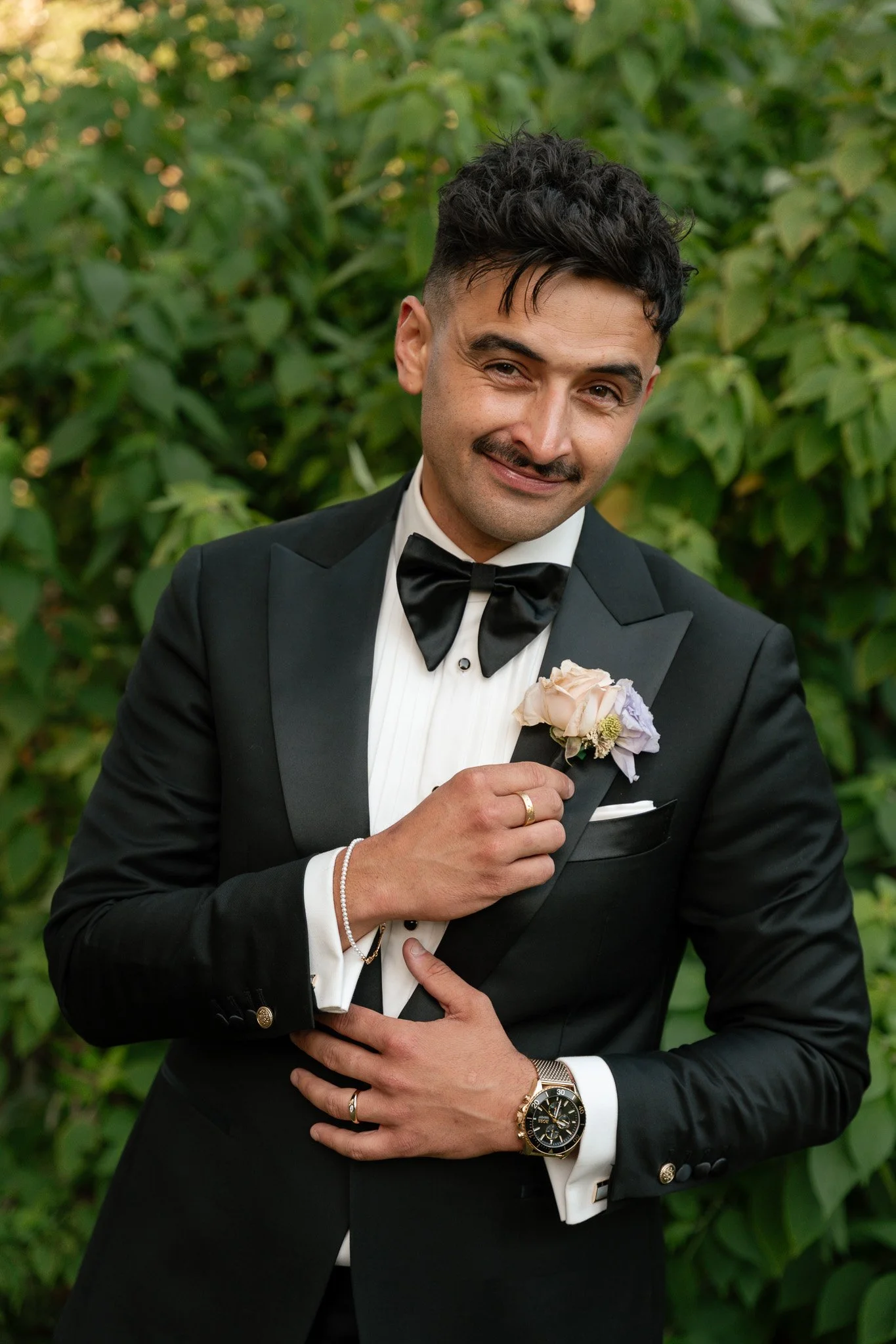 A man in a black tuxedo with a bow tie, wearing a wristwatch, rings, bracelets, and a boutonniere, is posing outdoors in front of green foliage.