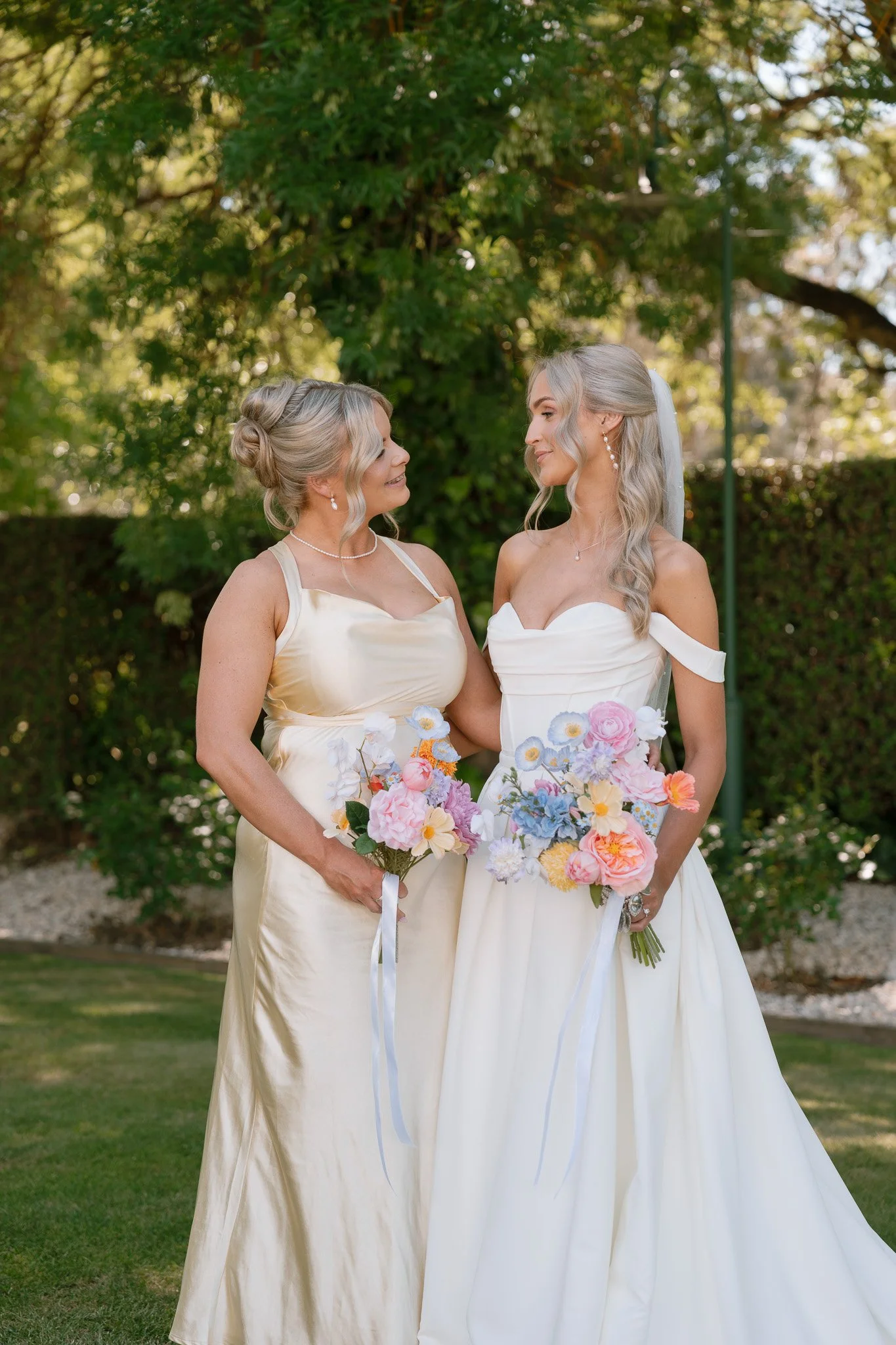 Two women in wedding dresses holding colorful bouquets standing outdoors with green trees in the background, smiling at each other.