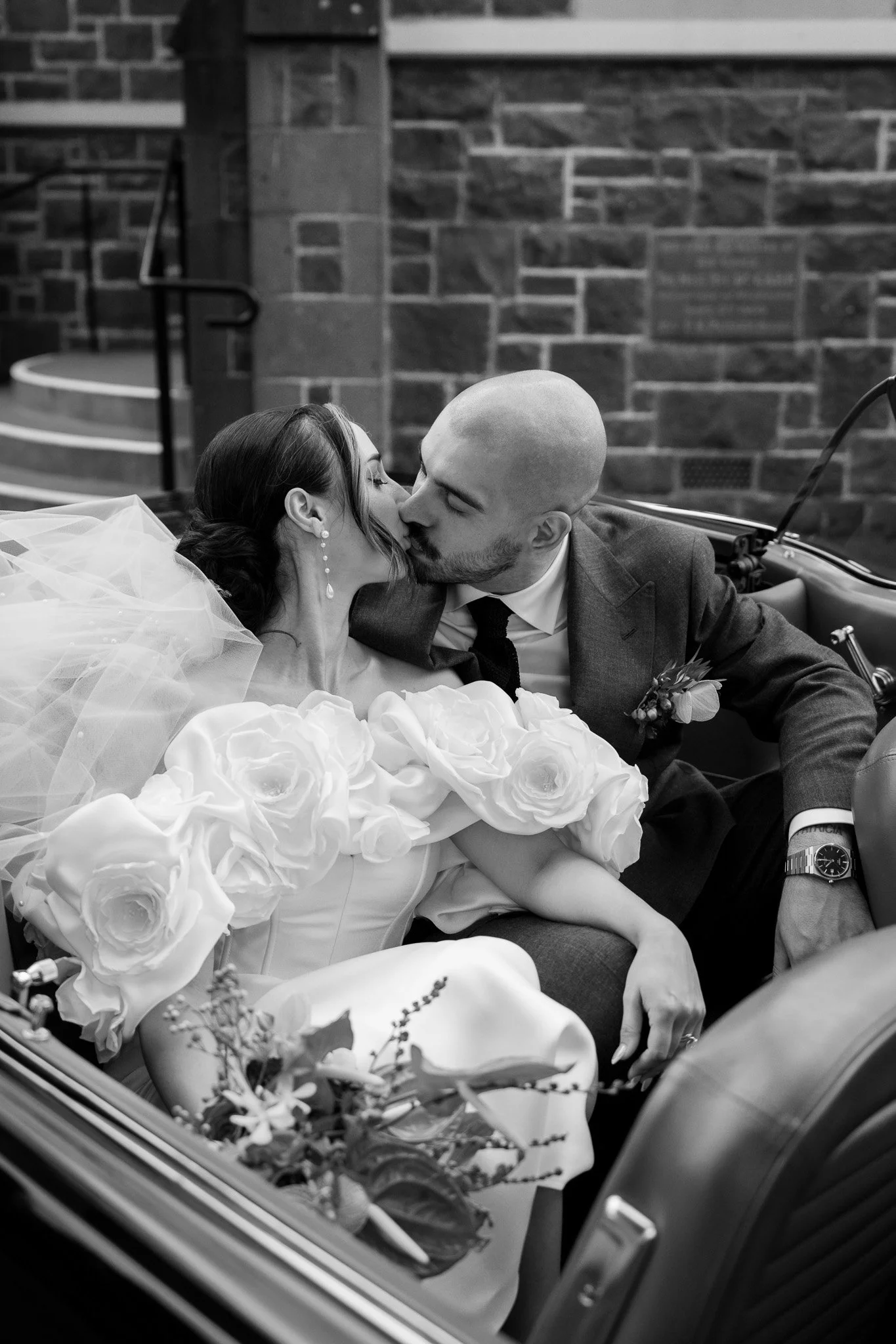 A bride and groom sharing a kiss in a vintage car, with the bride wearing a dress with large roses and the groom in a suit, against a brick wall.