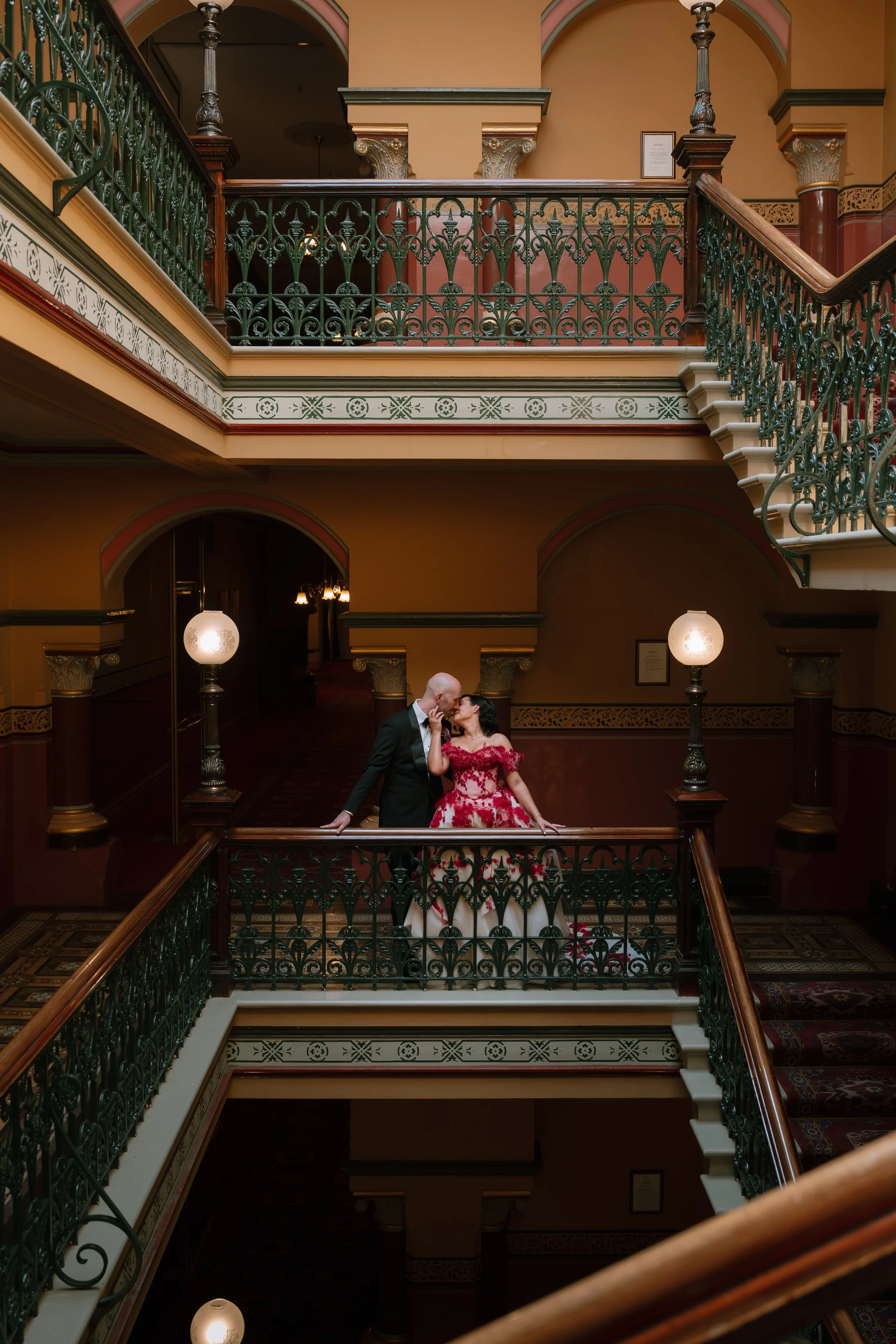A couple dressed in formal wedding attire standing on a decorative staircase in an elegant, vintage-style building, facing each other affectionately.