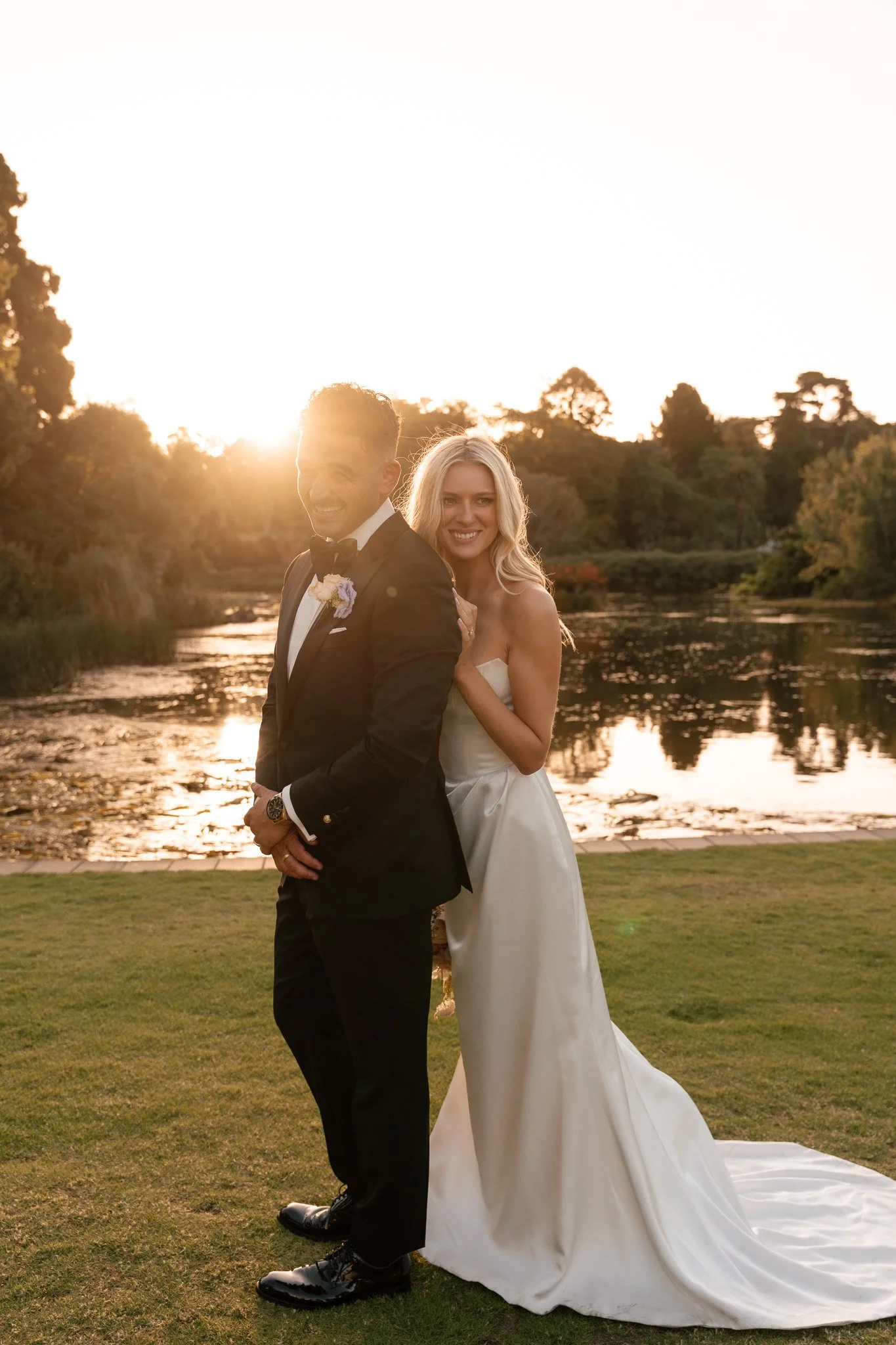A newlywed couple standing outside by a river at sunset, with trees and a grassy area in the background. The bride is wearing a strapless white wedding gown, and the groom is in a black tuxedo. They are smiling and embracing each other.