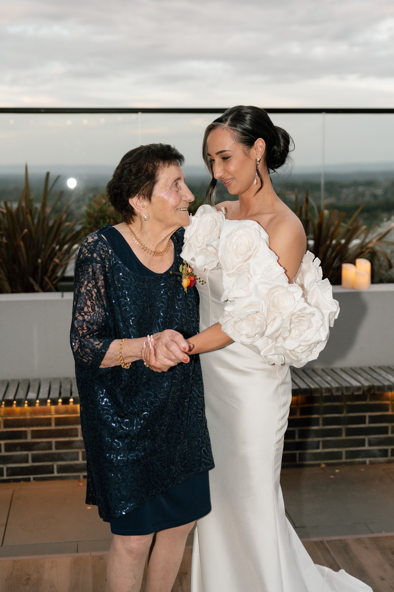 A bride in a white wedding dress with large floral details on her sleeves, dancing with an older woman in a navy blue dress with lace, on a rooftop with cityscape in the background.