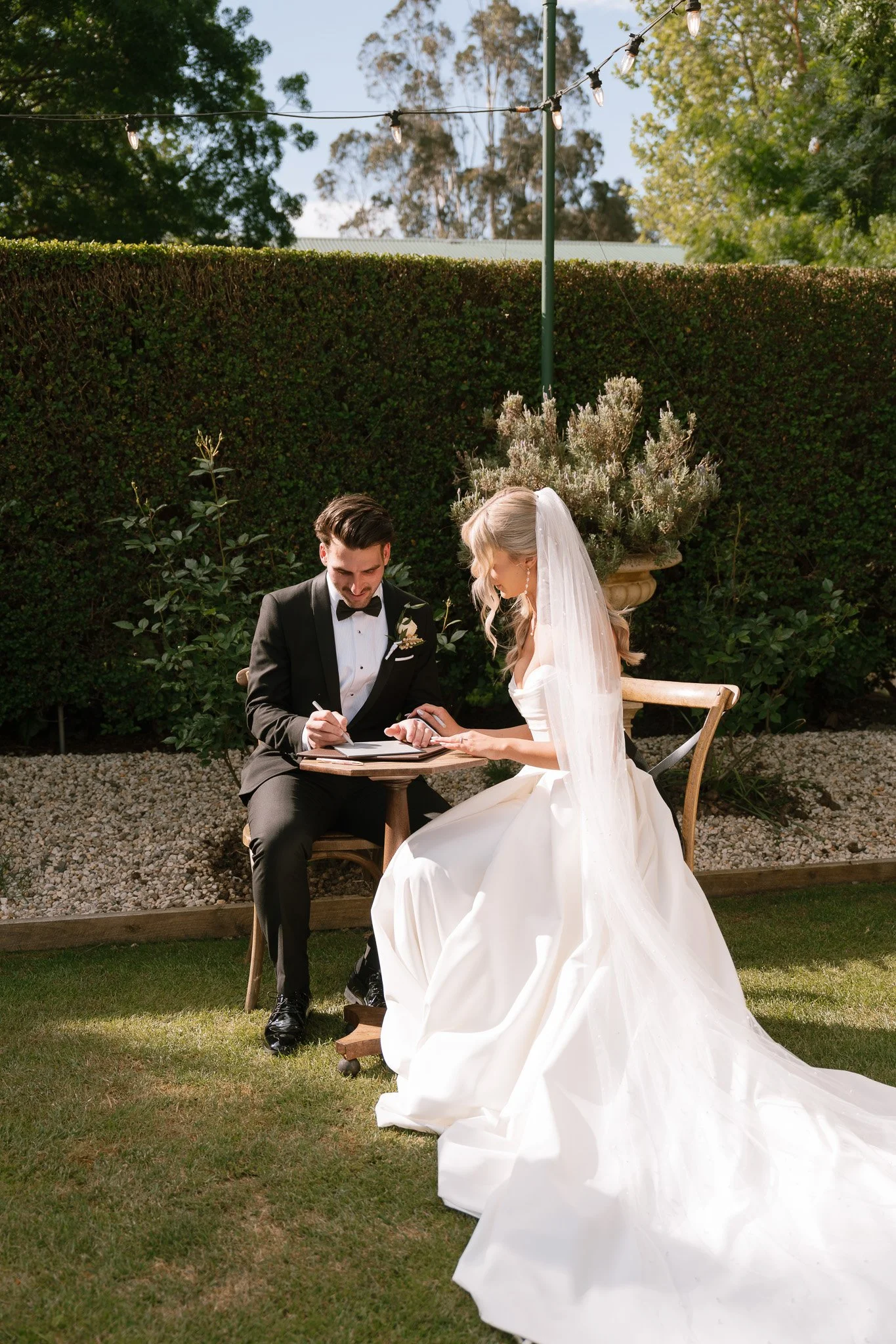 Bride and groom sit at a small outdoor table during their wedding ceremony, signing documents, on a sunny day with trees and string lights overhead.