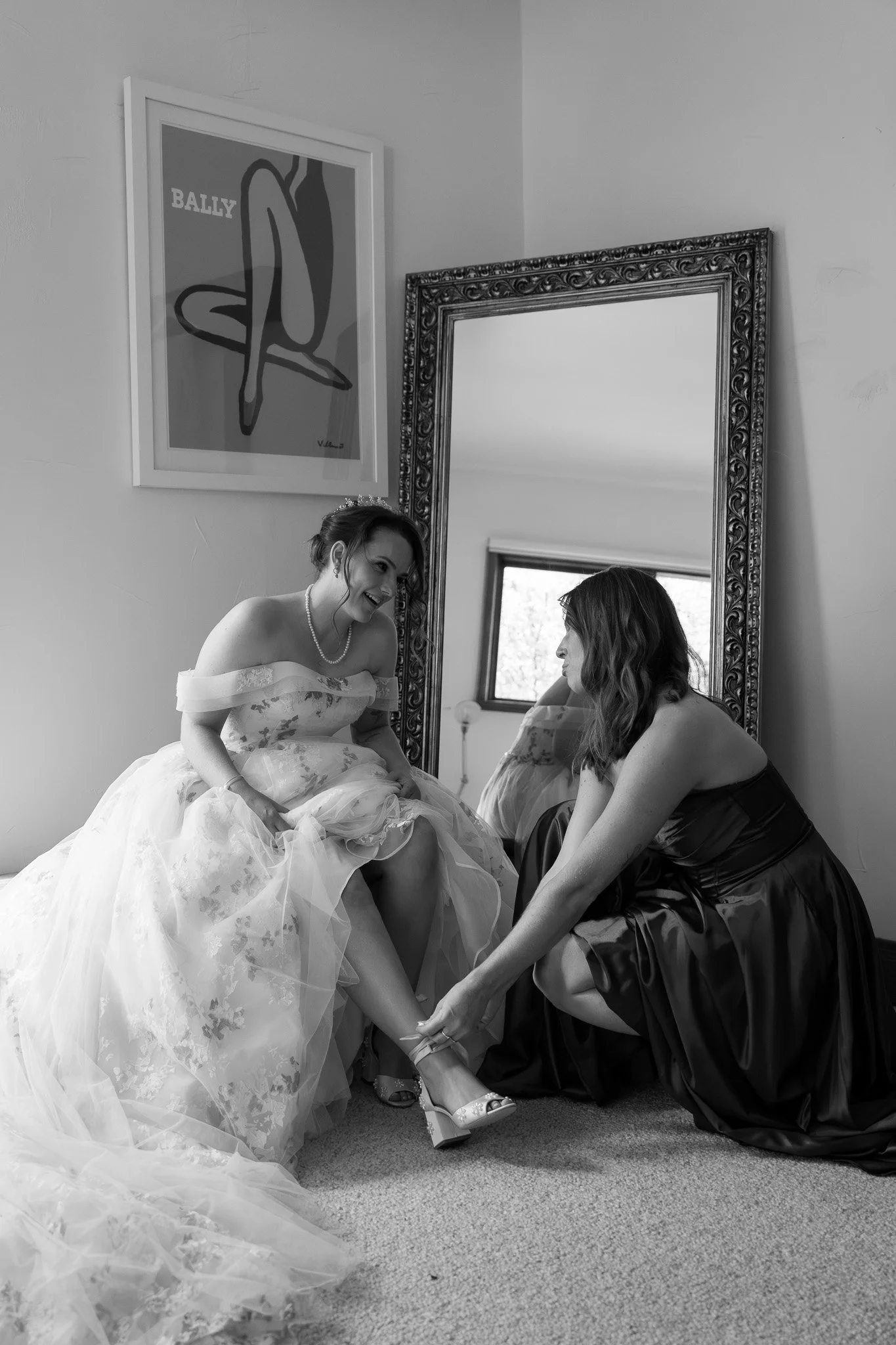 A bride in a wedding dress helping another woman get ready, as she puts on a shoe, in a room with a large mirror and a framed artwork on the wall.