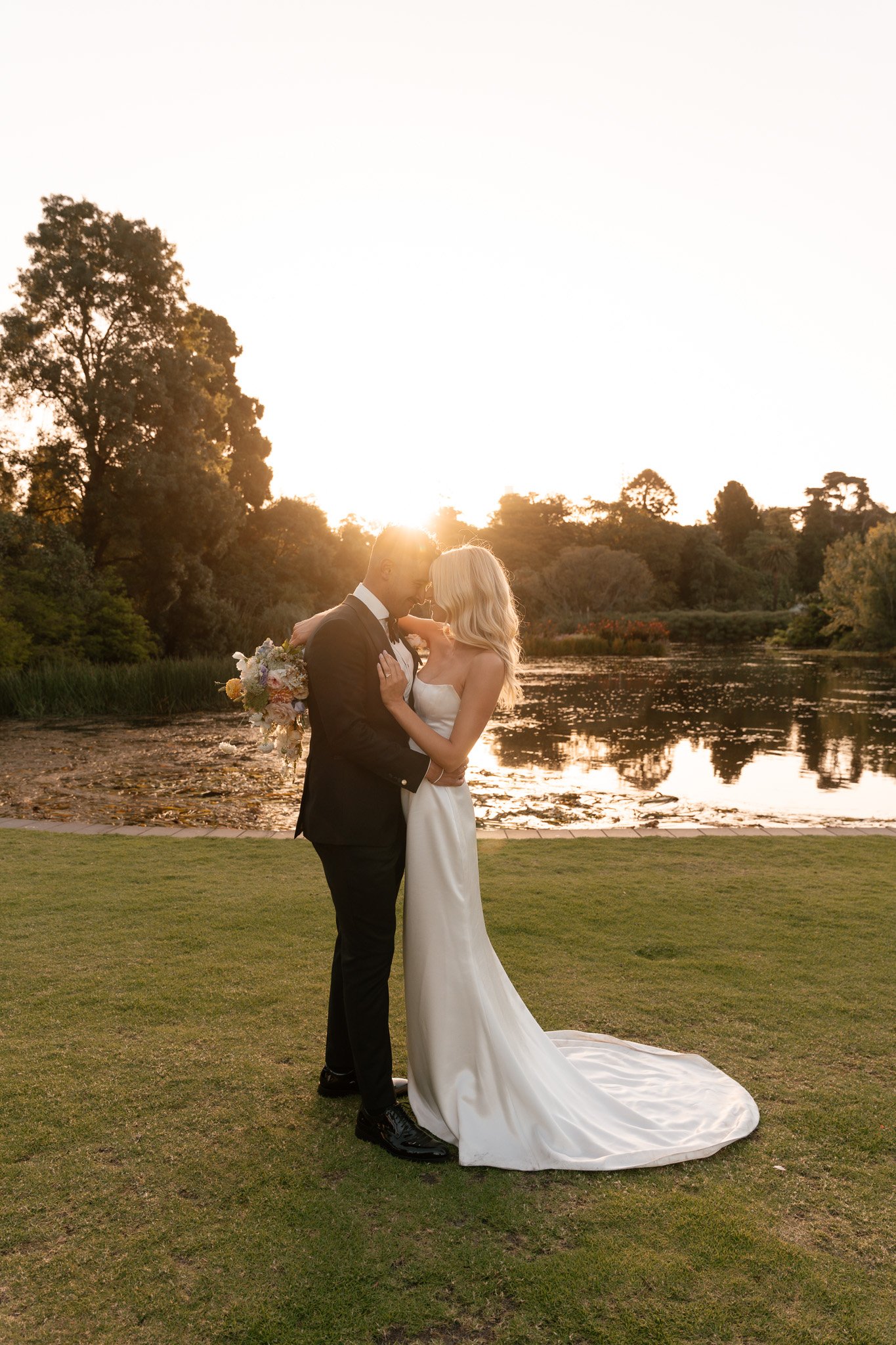 A bride and groom embrace during their wedding photos on a grassy area near a lake at sunset, with trees and water in the background.