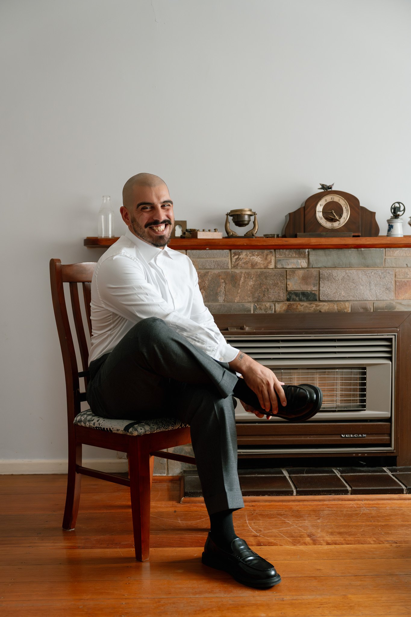 A man with a shaved head and beard, wearing a white dress shirt and gray trousers, sitting on a wooden chair, smiling and adjusting his black shoe in front of a fireplace with vintage decor.
