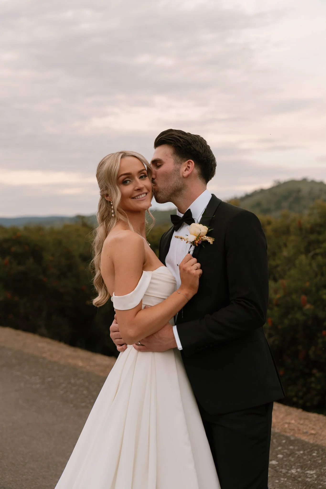 A newlywed couple dressed in wedding attire sharing an intimate moment outdoors. The bride is wearing a strapless white gown with off-the-shoulder sleeves, and the groom is in a black tuxedo with a bow tie. The groom is kissing the bride on the cheek