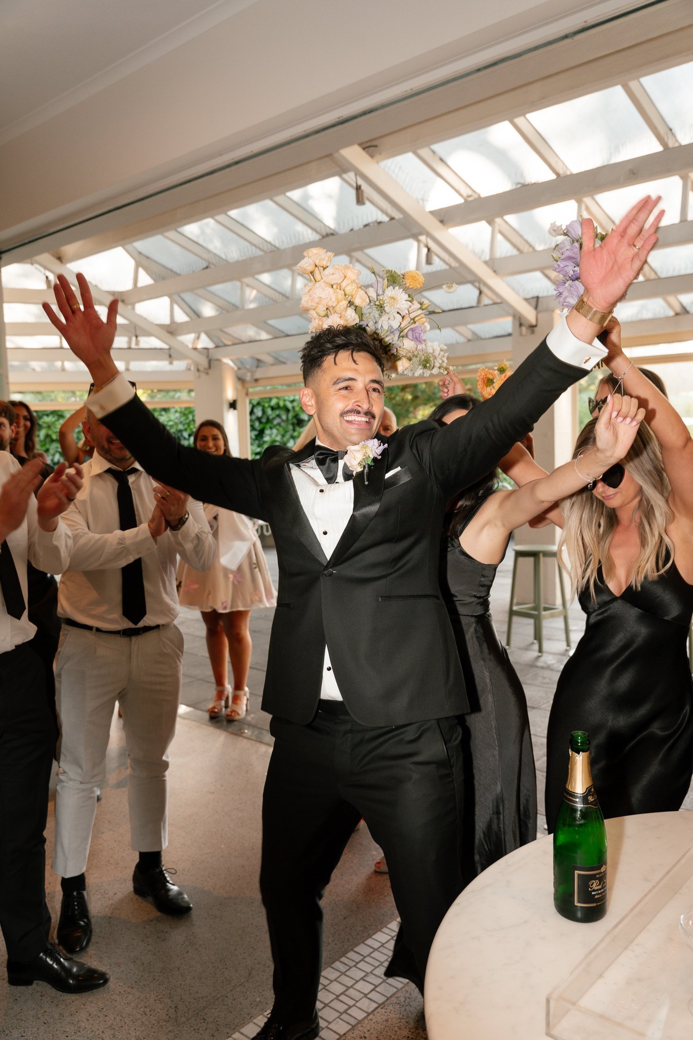 A man in a tuxedo with a boutonniere raises his hands in celebration at a wedding reception, surrounded by guests clapping and smiling.