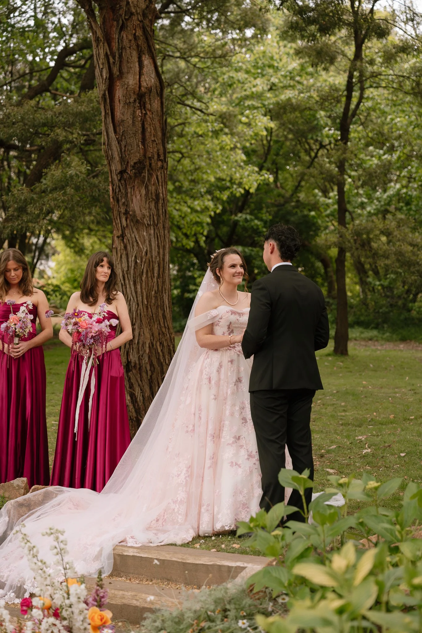 Wedding ceremony outdoors with bride in a pink gown and veil, groom in a black tuxedo, and two bridesmaids in pink dresses holding bouquets, standing on a grassy area near a large tree.
