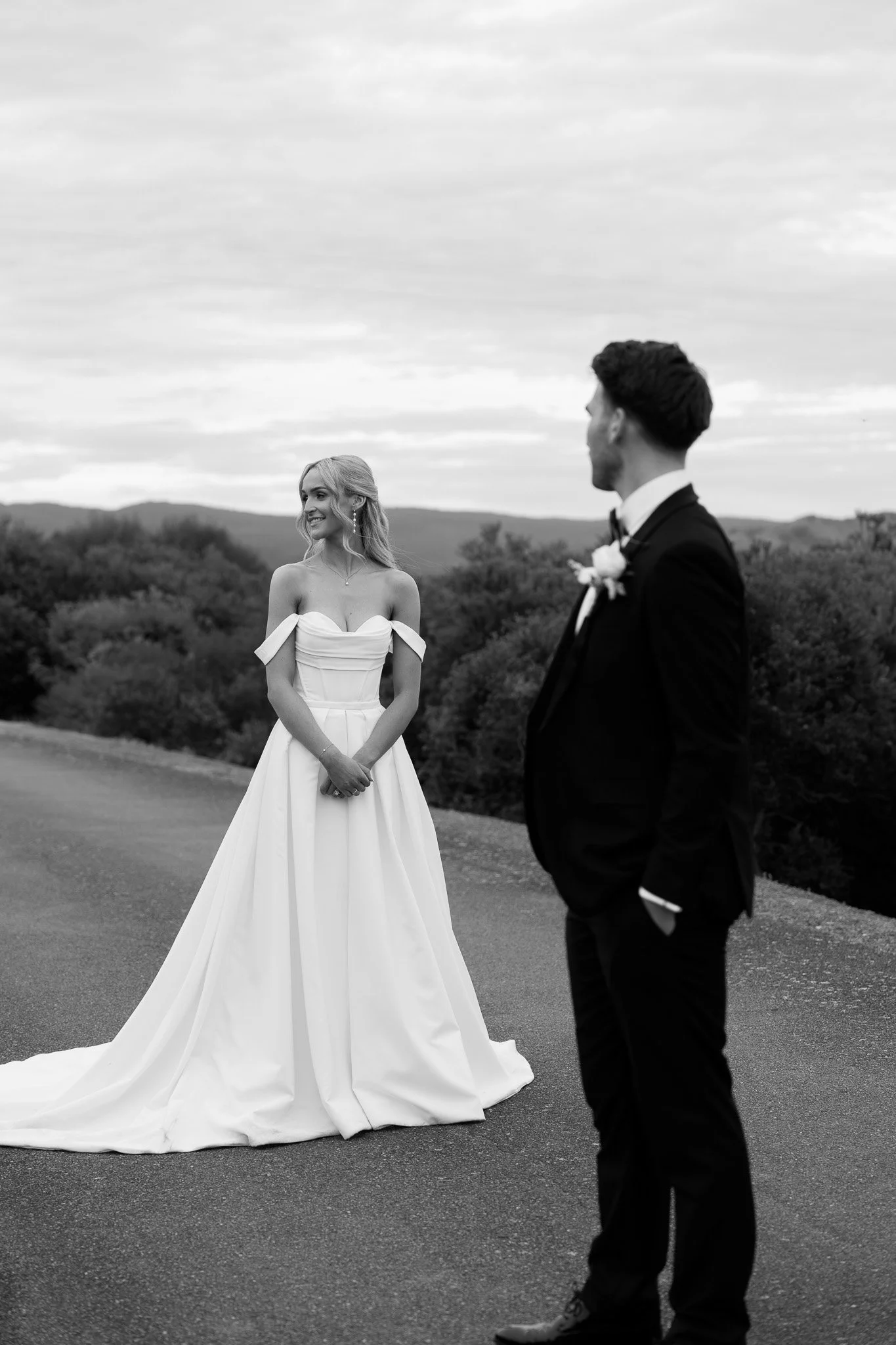A bride in a wedding gown and a groom in a suit standing outdoors on a road with trees and mountains in the background.