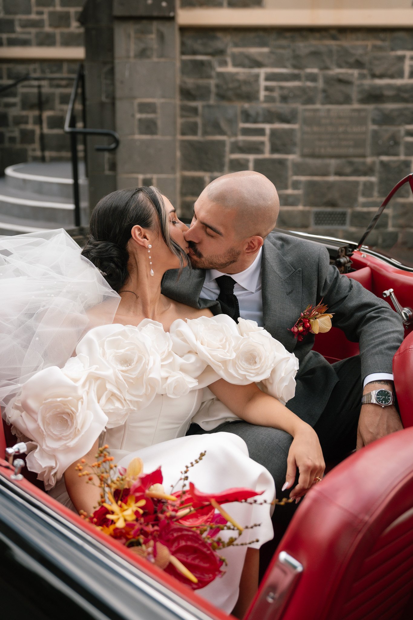 A bride and groom sharing a kiss in a vintage red car, with the bride wearing a white dress with large floral details and the groom in a grey suit and black tie, against a stone building background.