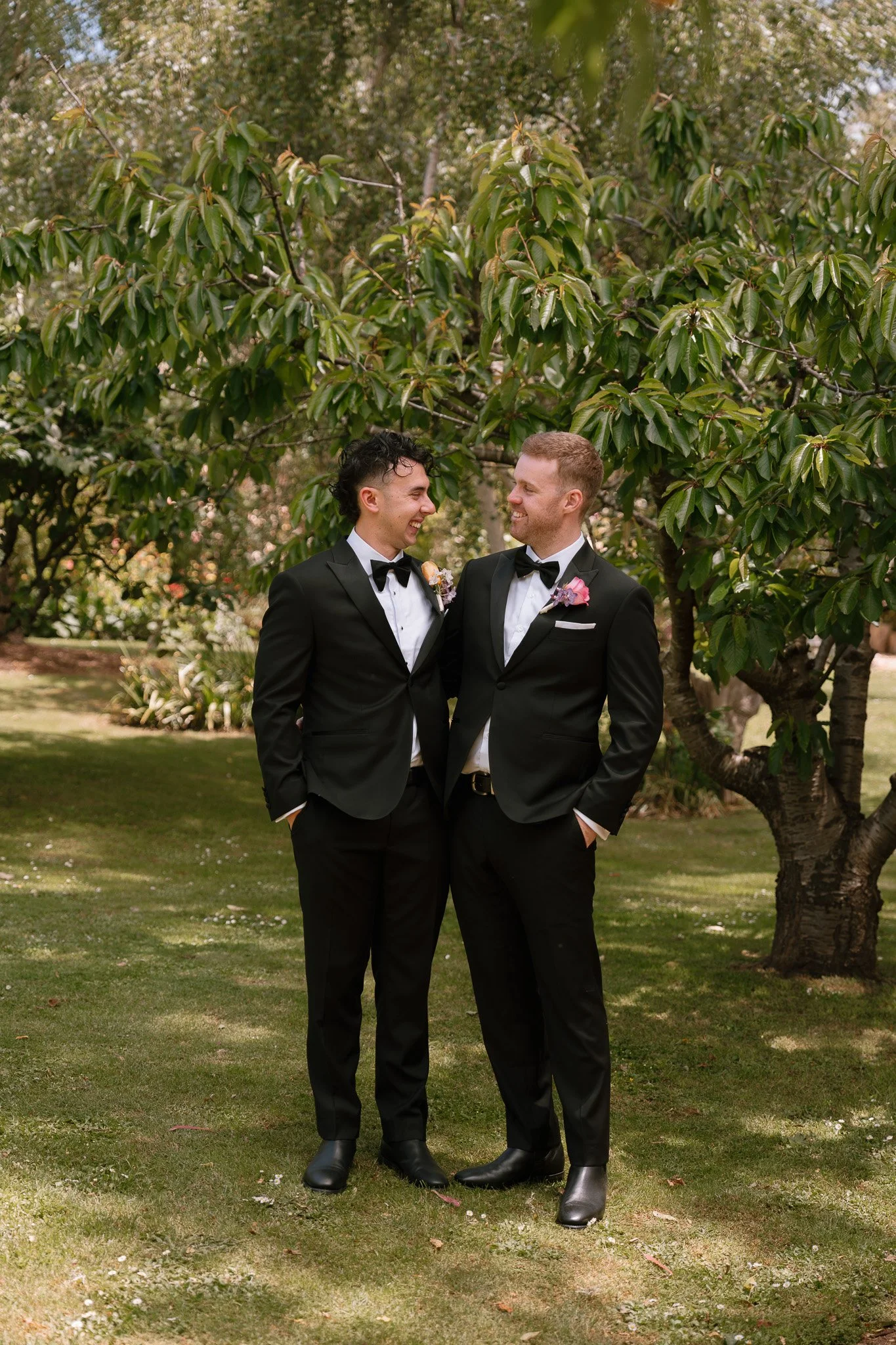 Two men in tuxedos smiling and looking at each other outdoors under a tree.
