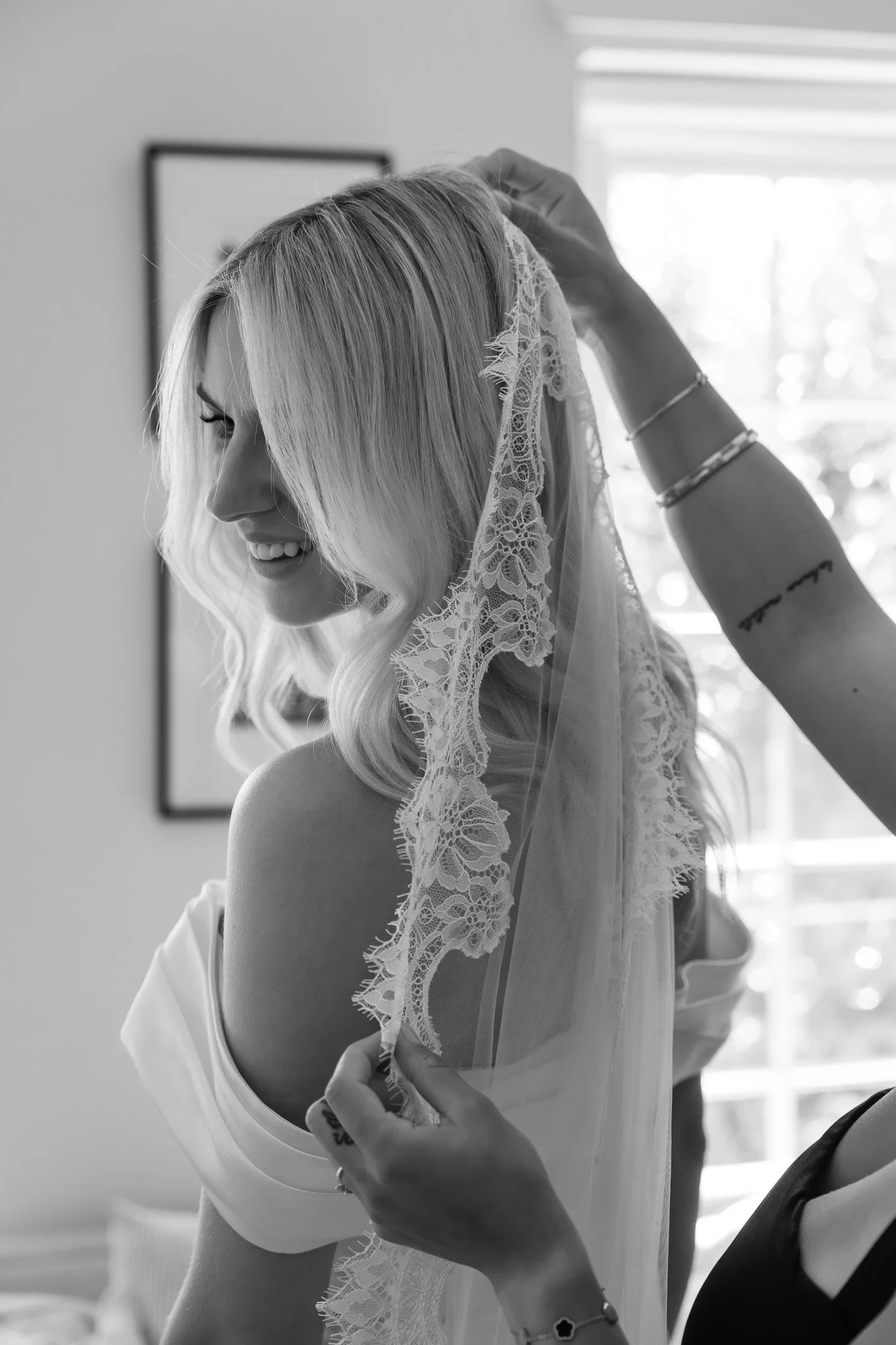 A woman with blonde hair smiling as someone adjusts a lace veil on her head in a bright room.