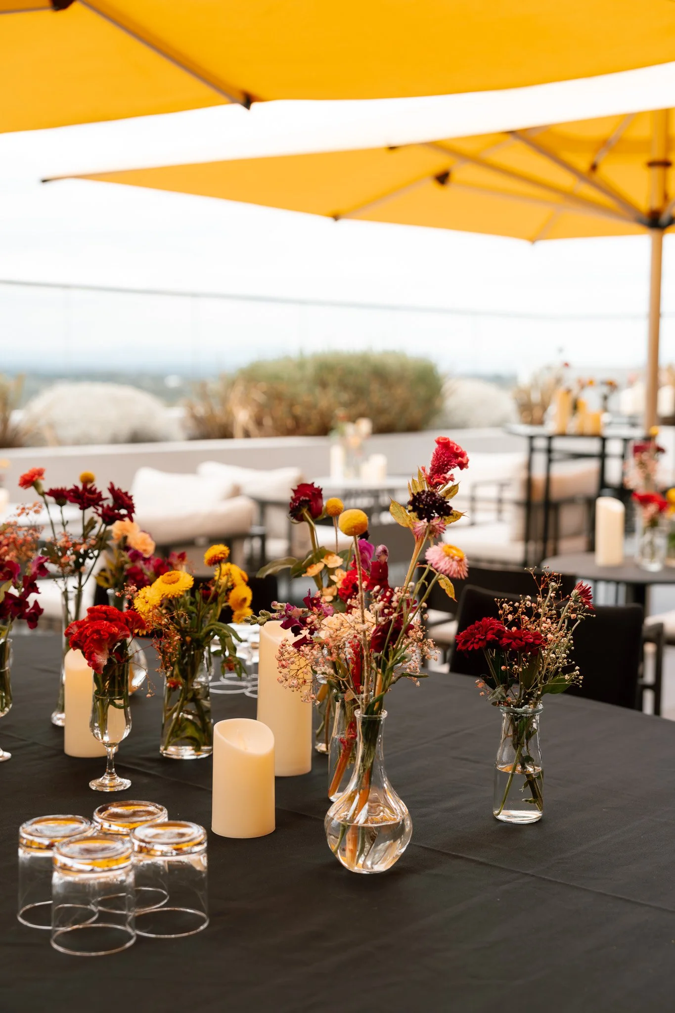 A dining setup on a black table with floral arrangements, candles, and glassware under a yellow umbrella on a patio.
