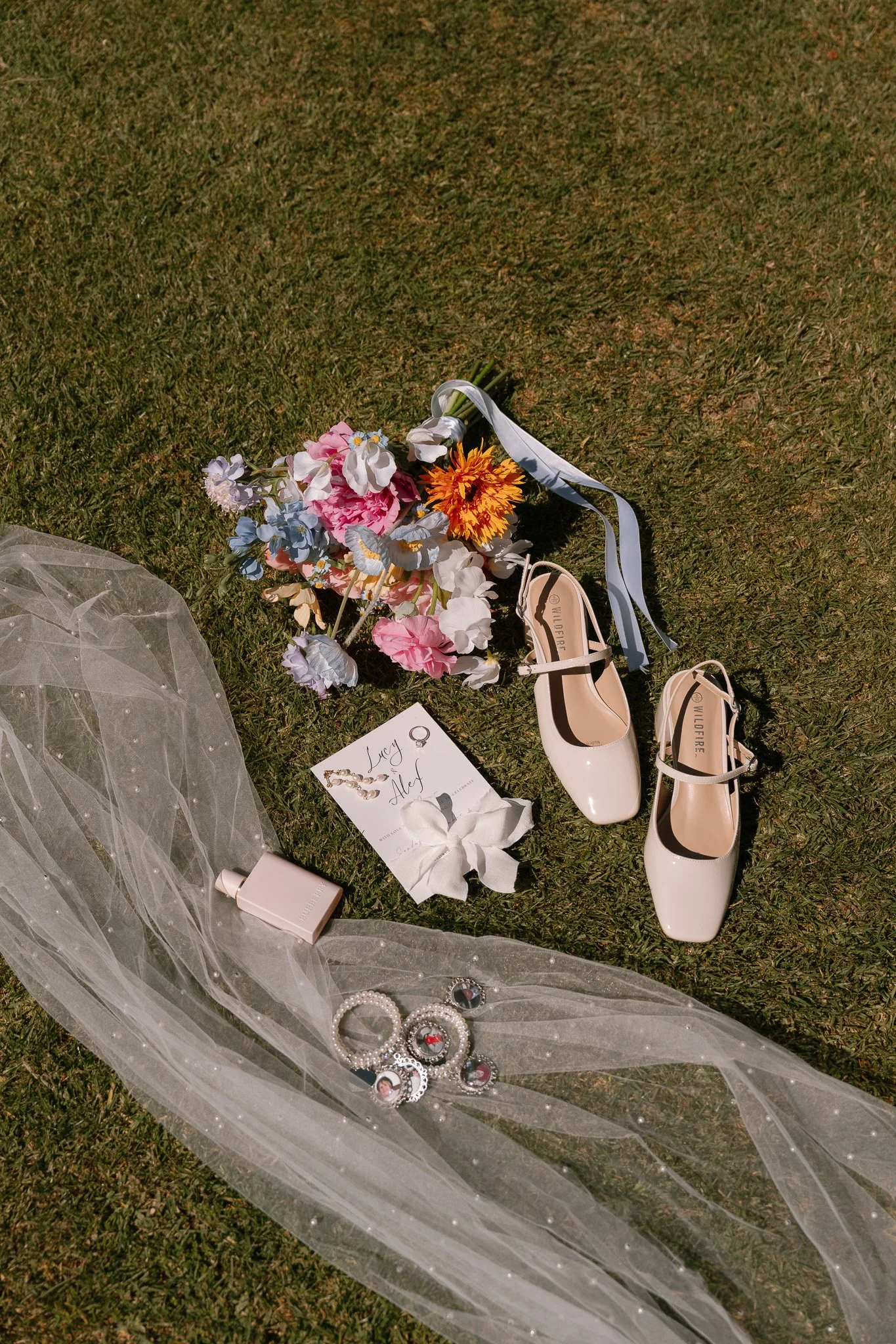 Wedding accessories and decorations on grass, including a bouquet of flowers, white shoes, a veil with pearls, jewelry, perfume, and a card.