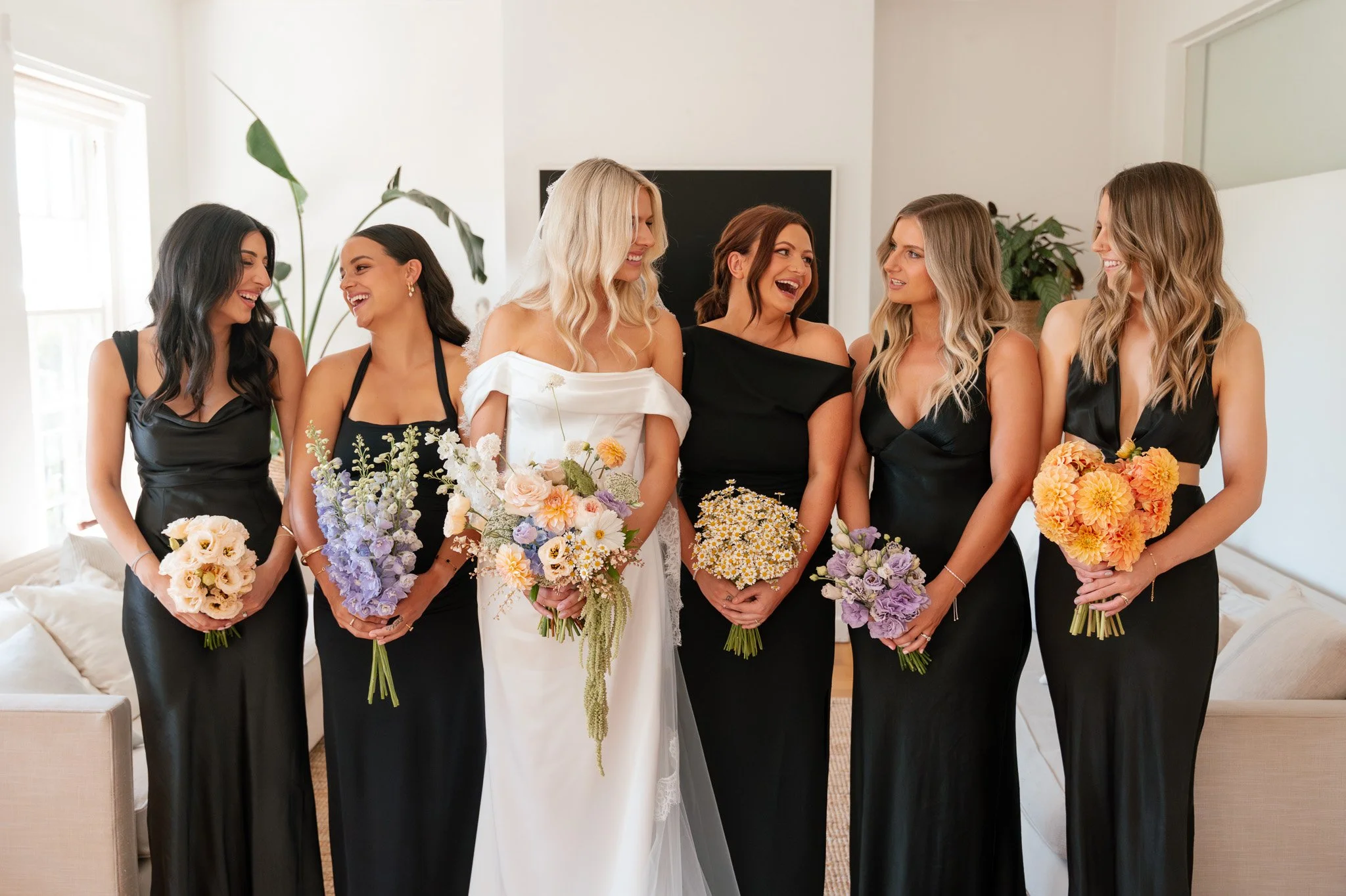 A bride and five bridesmaids standing indoors, all holding bouquets of flowers, smiling and engaging in conversation.