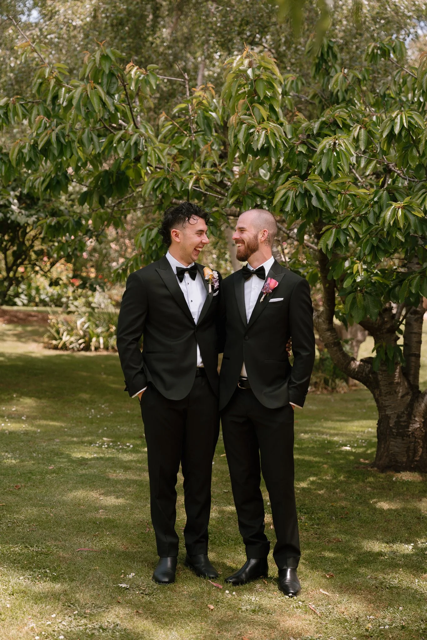 Two men in tuxedos standing on grass in front of a tree, smiling and looking at each other, celebrating a wedding.