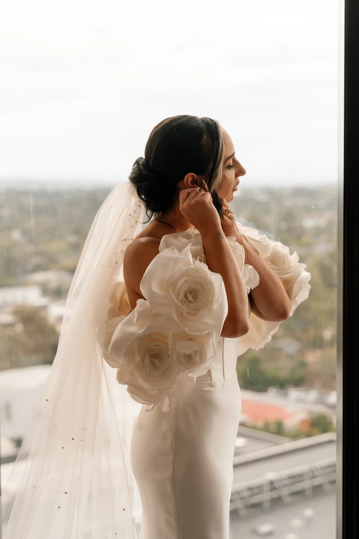 A bride with dark hair styled in an updo, wearing a strapless white wedding dress with large 3D floral embellishments on the sleeves. She is adjusting her earrings while standing near a window, overlooking a cityscape on an overcast day.