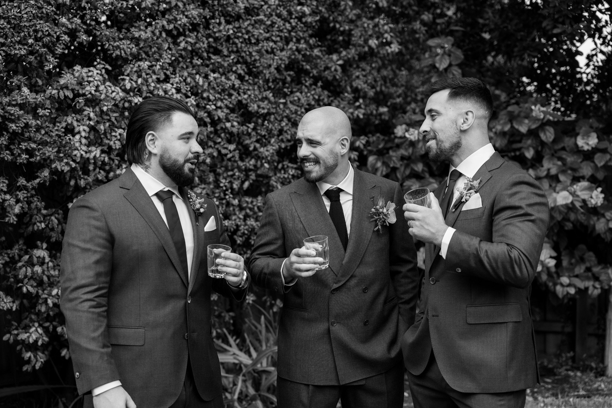 Three men in suits and ties, holding glasses, talking and smiling outdoors with a bush in the background.